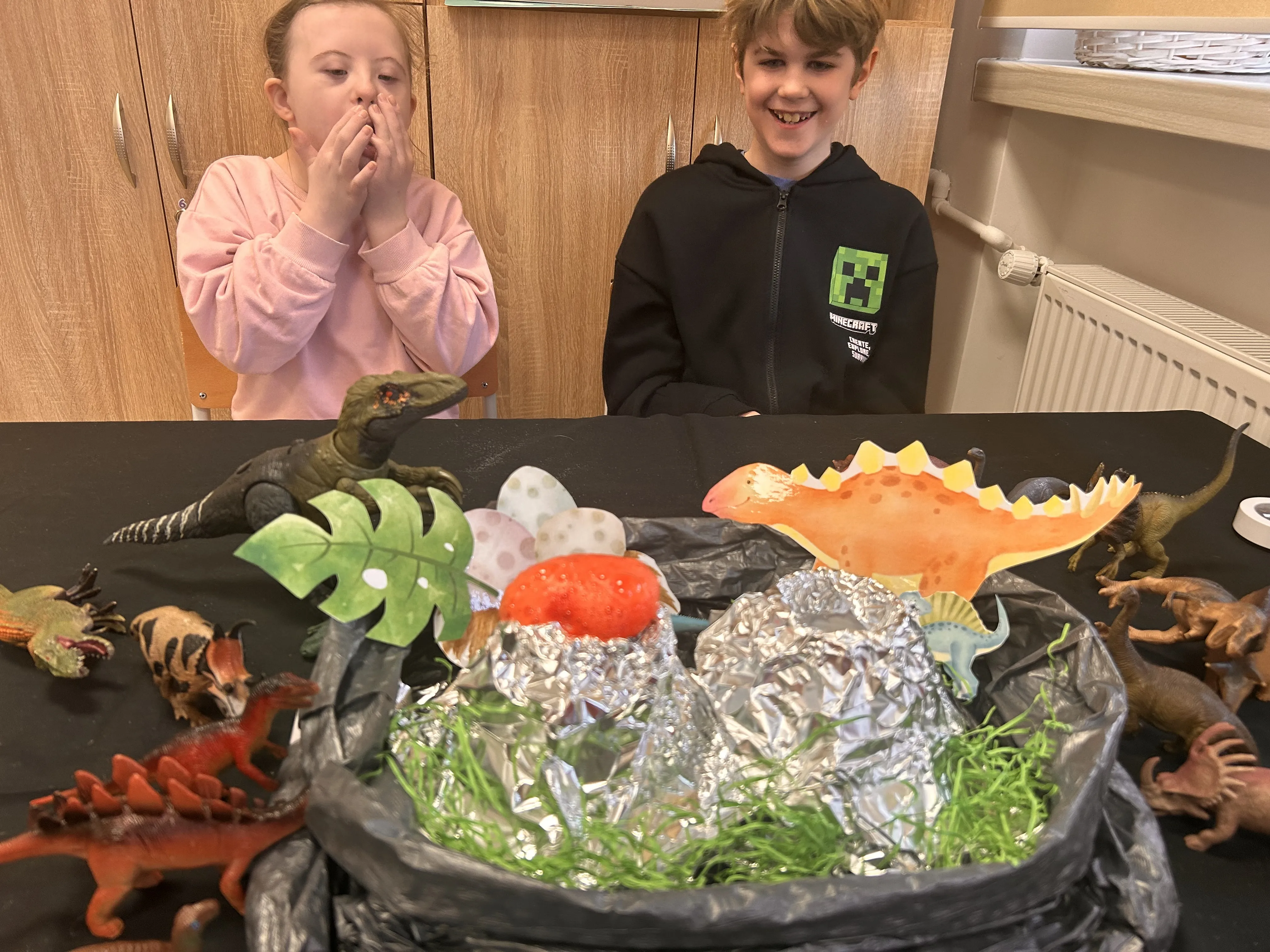 Na zdjęciu uczeń i uczennica przy stoliku, na którym znajdują się różne modele dinozaurów oraz model wulkanu