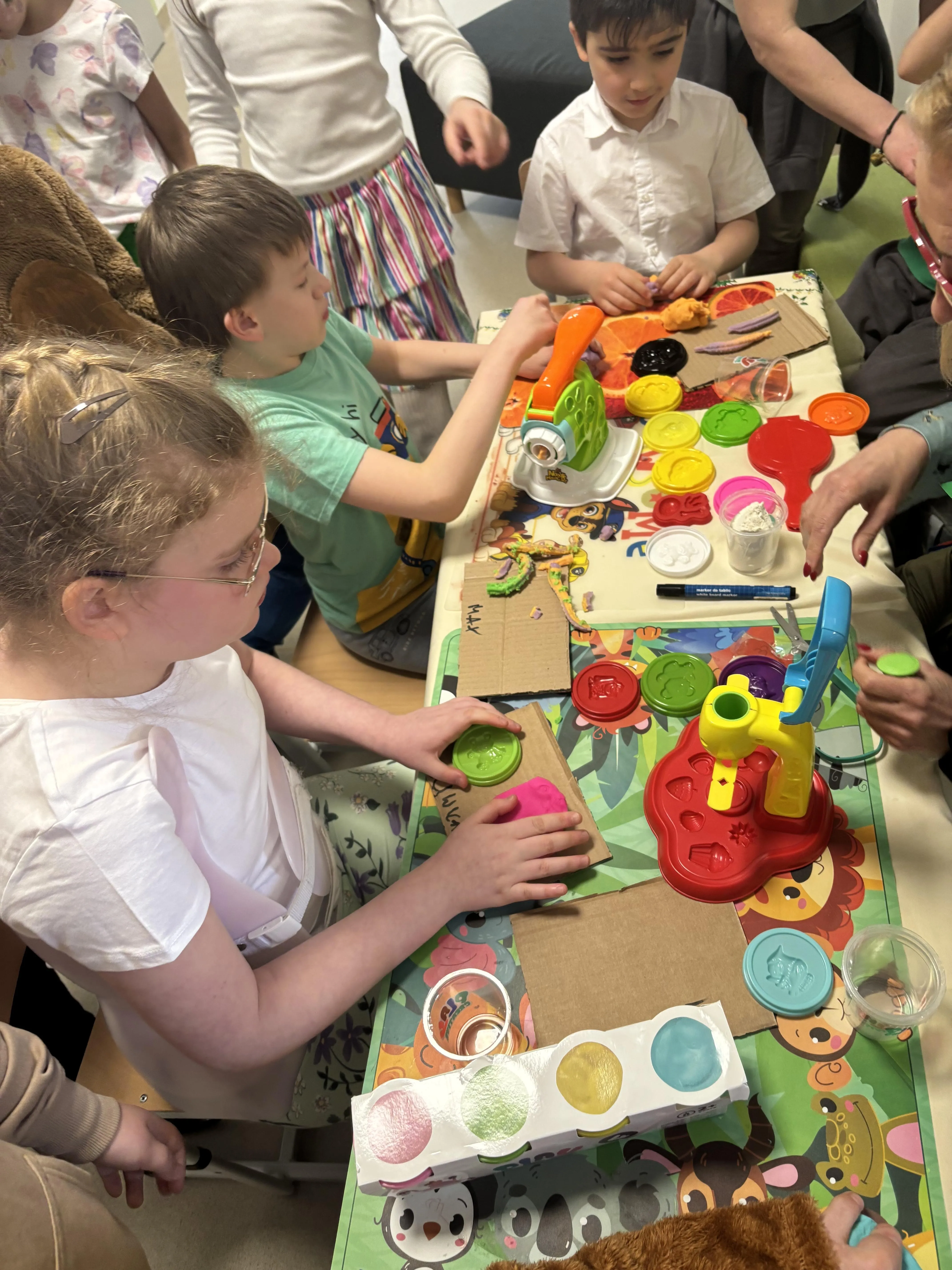 Przy dużym podłużnym stole, na którym rozłożone są różne pomoce   siedzą uczniowie, którzy zajmują się układaniem, lepieniem, wycinaniem 