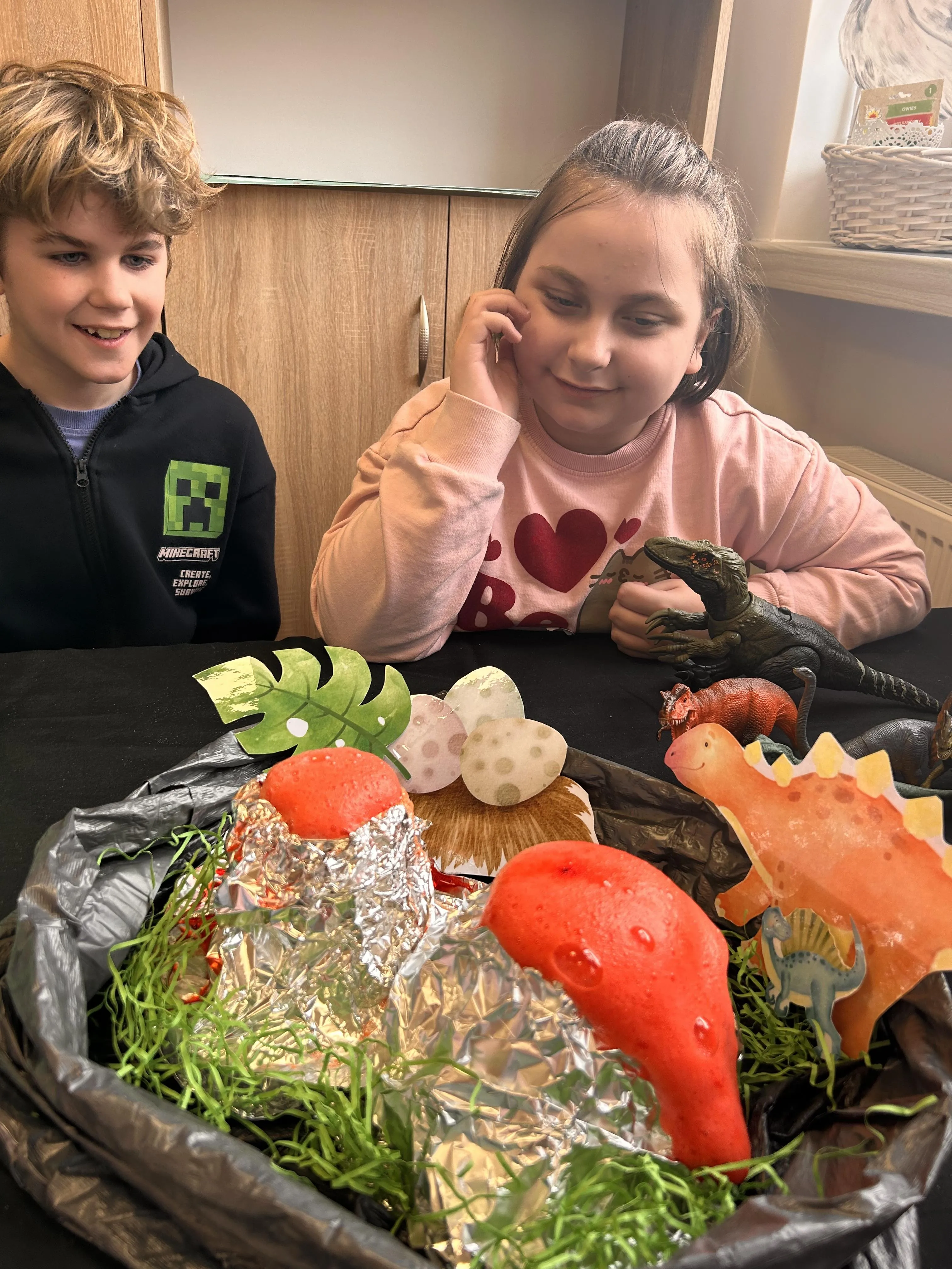 Dwoje uczniów siedzi przy stoliku, na którym ułożone są  akcesoria związane z życiem dinozaurów