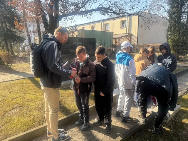 Sześcioro uczniów z dwojgiem nauczycieli na cmentarzu Powstańców Warszawskich - na pierwszym planie  nauczyciel pomaga zapalić znicz uczniowi, dalej  stoi grupa uczniów wraz  z nauczycielką, jeden z uczniów wyjmuje z torby znicze.