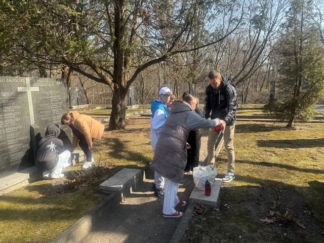 Uczniowie z dwojgiem nauczycieli na cmentarzu Powstańców Warszawskich - dwóch uczniów  pochylonych jest przy jednej z mogił, opodal  nauczyciele wyjmują  z torby przyniesione znicze, koło nich stoją inni uczniowie. uczniowie. 