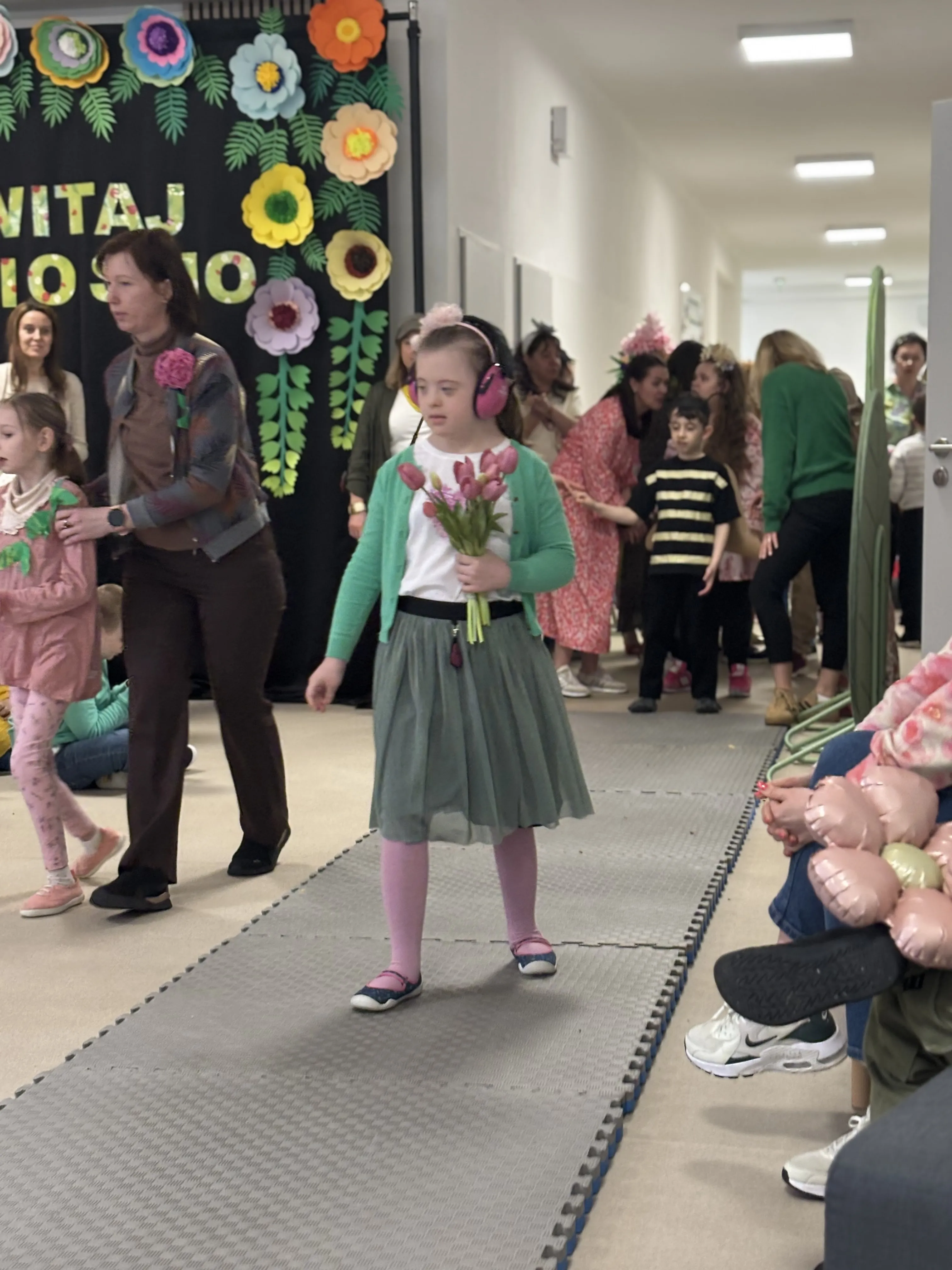 Pokaz mody: po rozłożonym dywanie idzie uczennica z tulipanami w ręku. Za nią widoczni są inni przygotowujący się do   przejścia uczniowie. W tle widoczna jest również  tablica z dekoracjami  wiosennymi