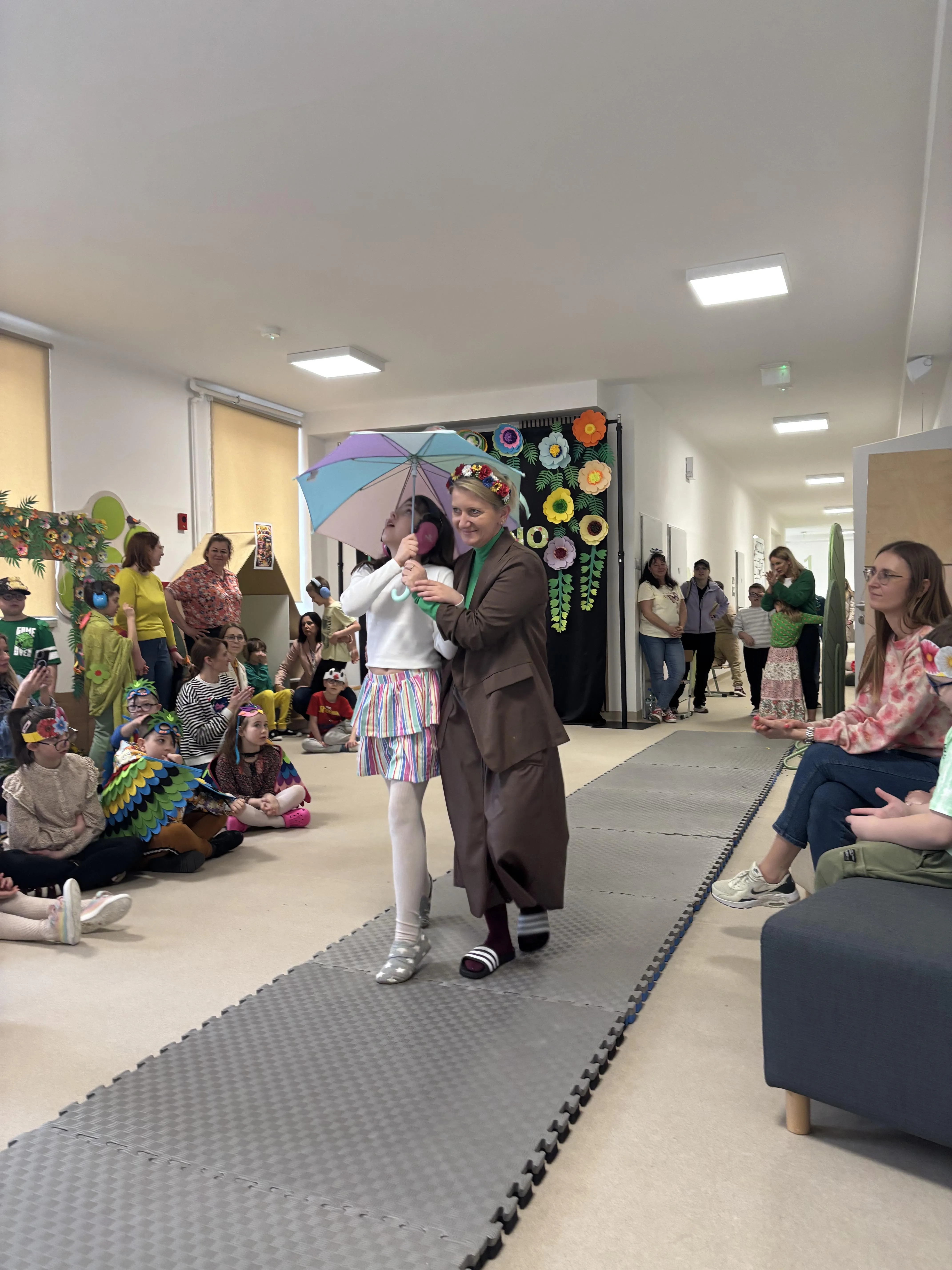 Pokaz mody:  po dywanie maszeruje uczennica z panią  - pomocą nauczyciela. W tle siedzący inni uczniowie .