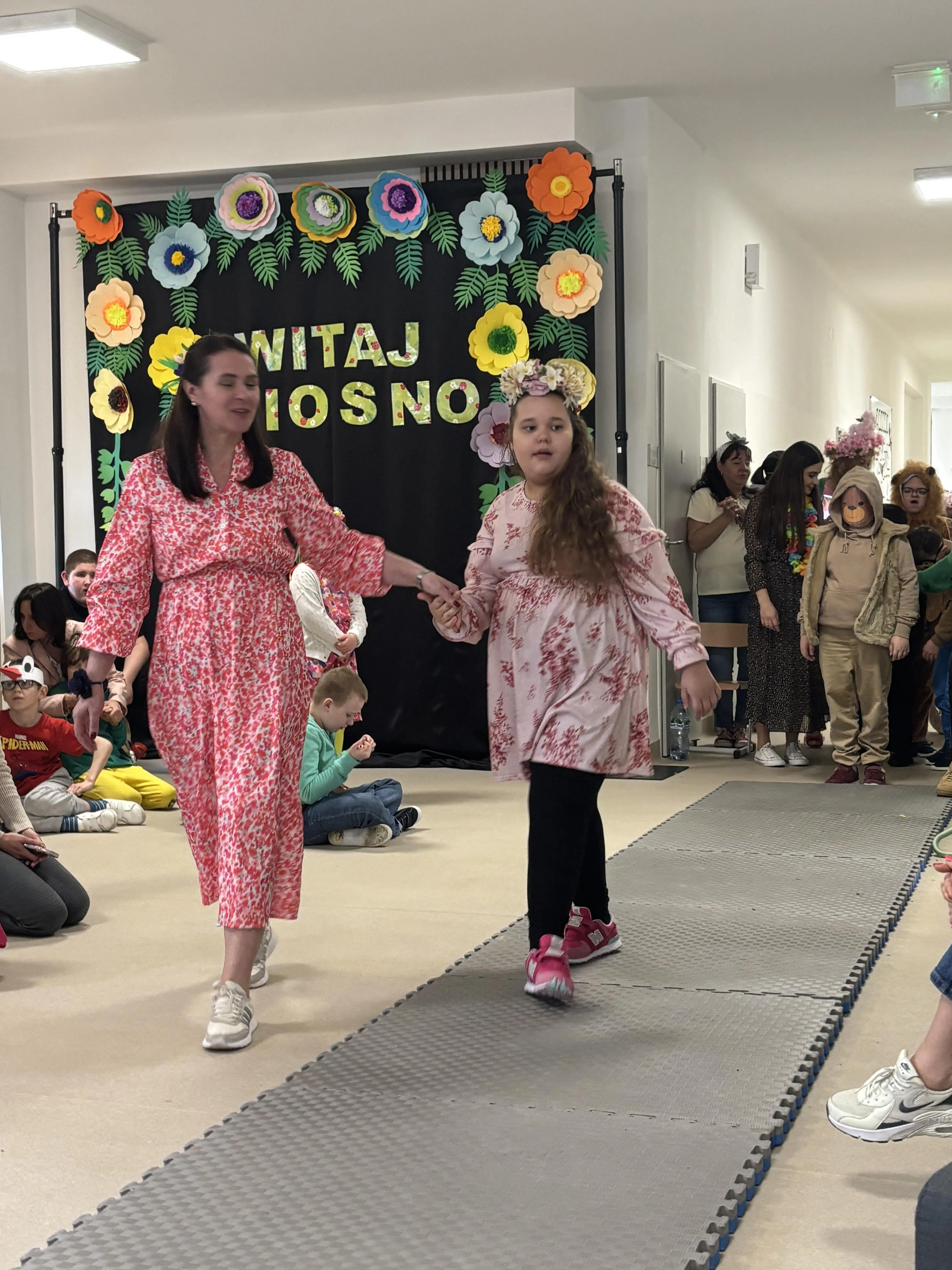 Pokaz mody: po rozłożonym dywanie idzie uczennica z panią nauczycielką.  Za nią  widoczni są inni przygotowujący się do przejścia. 