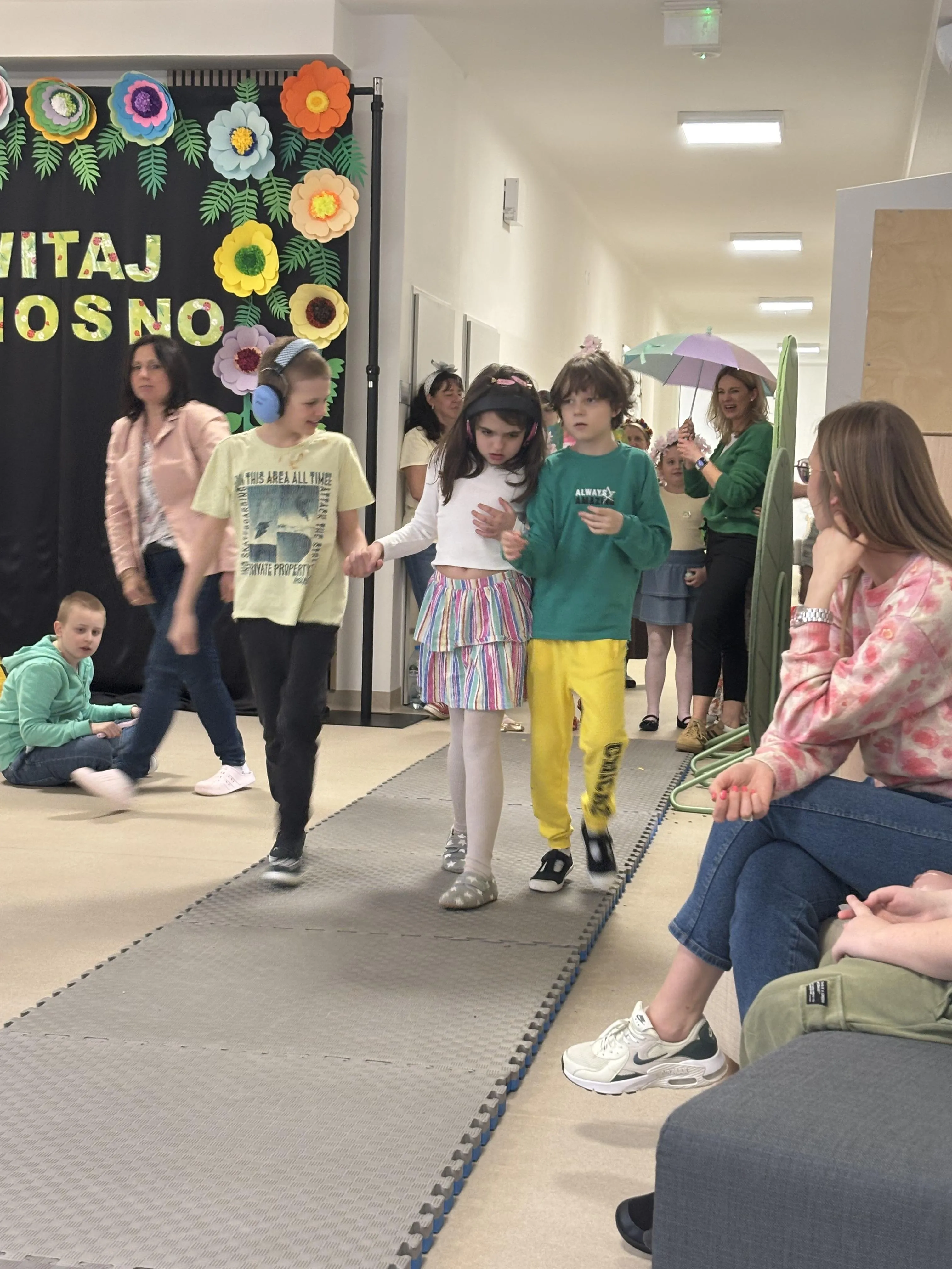 Pokaz mody: po rozłożonym dywanie idzie troje uczniów obok widoczna jest pani wychowawczyni. Za nimi widoczni są inni uczniowie z nauczycielami. Z boku widoczna jedna z pań nauczycielek obserwująca pokaz.