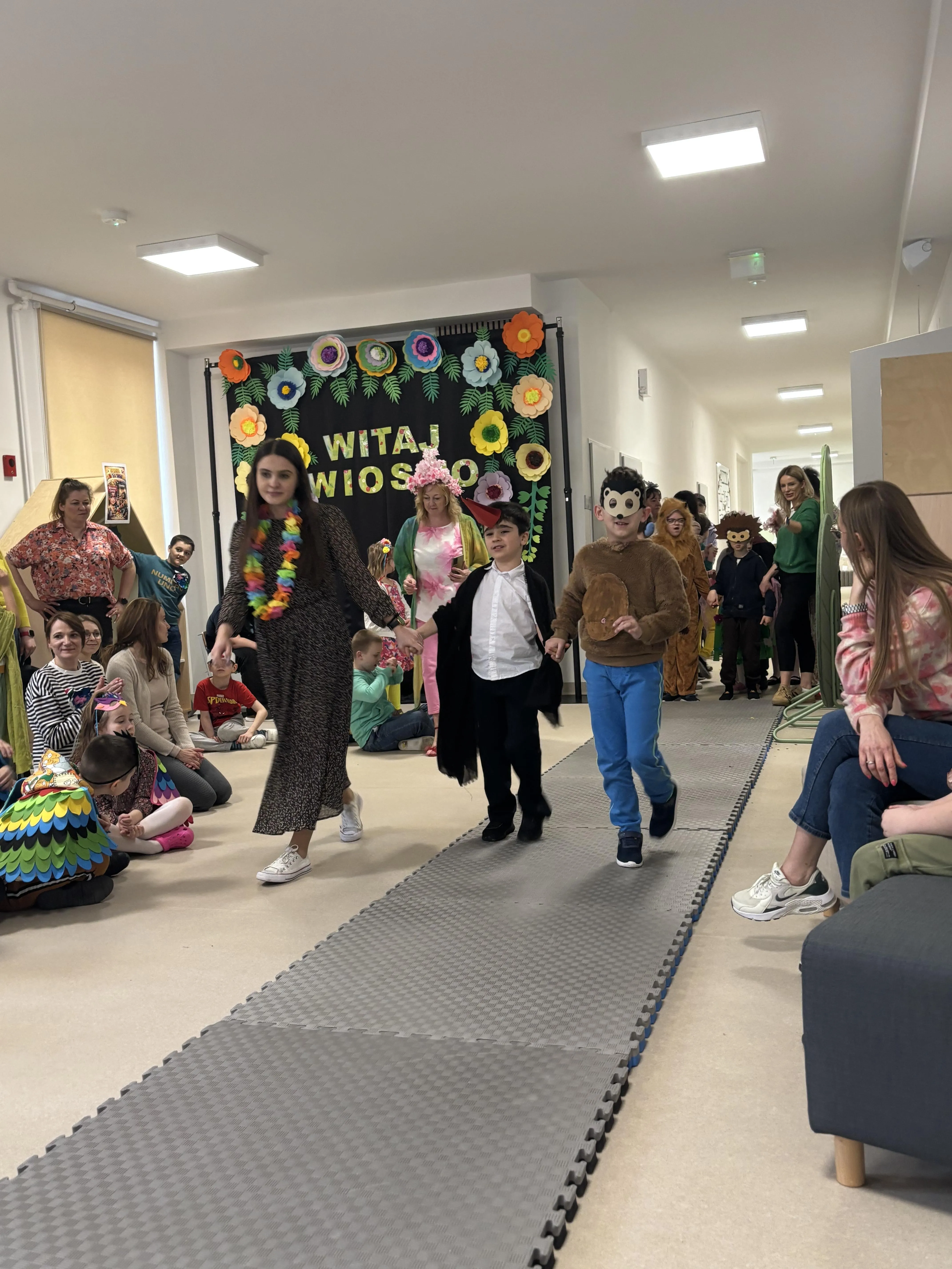 Pokaz mody: po rozłożonym dywanie idzie dwóch uczniów z panią nauczycielką,. Po obu stronach dywanu obserwują ich inni uczniowie oraz nauczyciele. W tle widać innych , czekających na swoje wyjście uczniów.