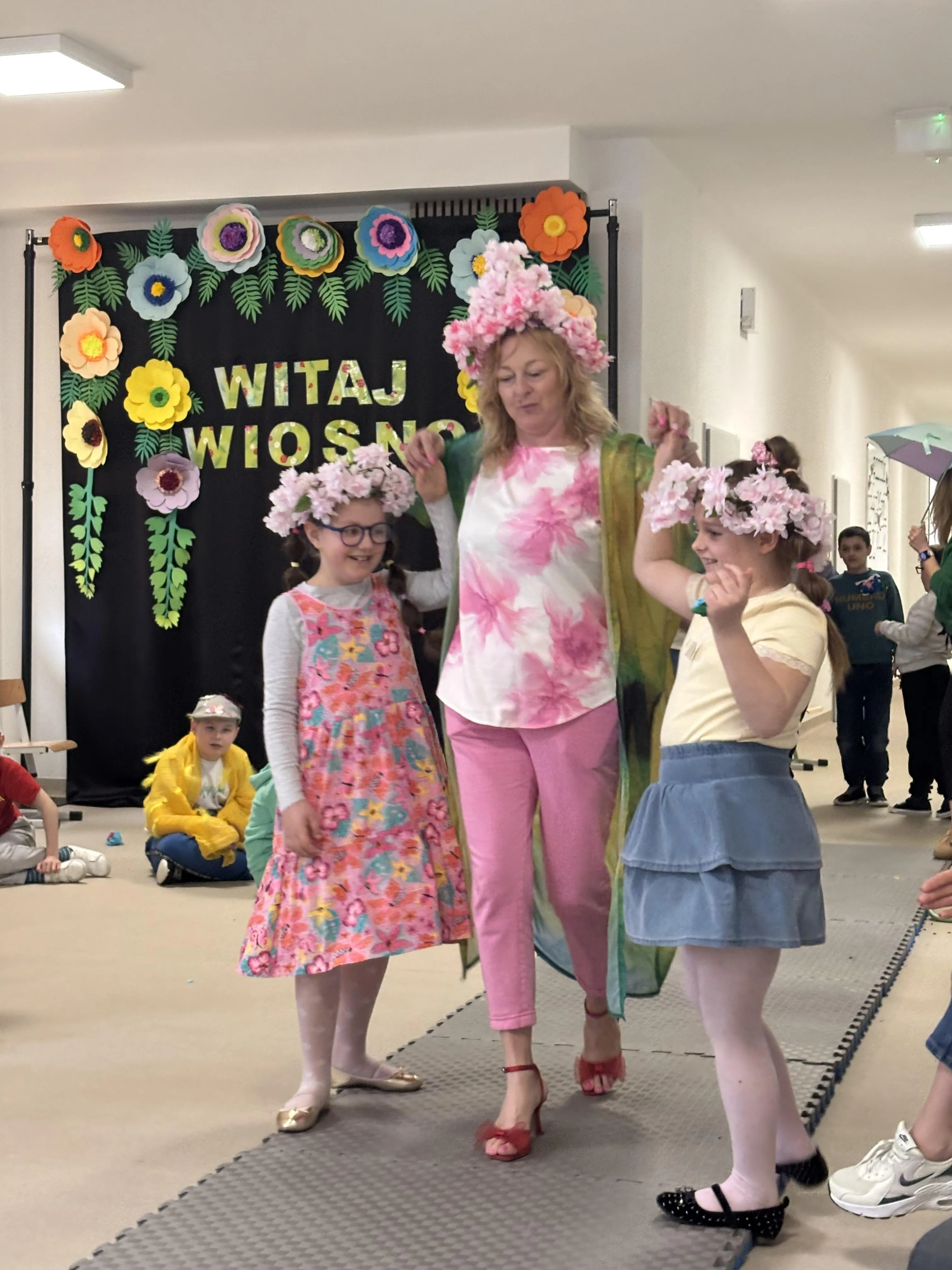 Pokaz mody: po rozłożonym dywanie idą dwie uczennice z wychowawczynią.  W tle widoczne są wiosenne kolorowe dekoracje