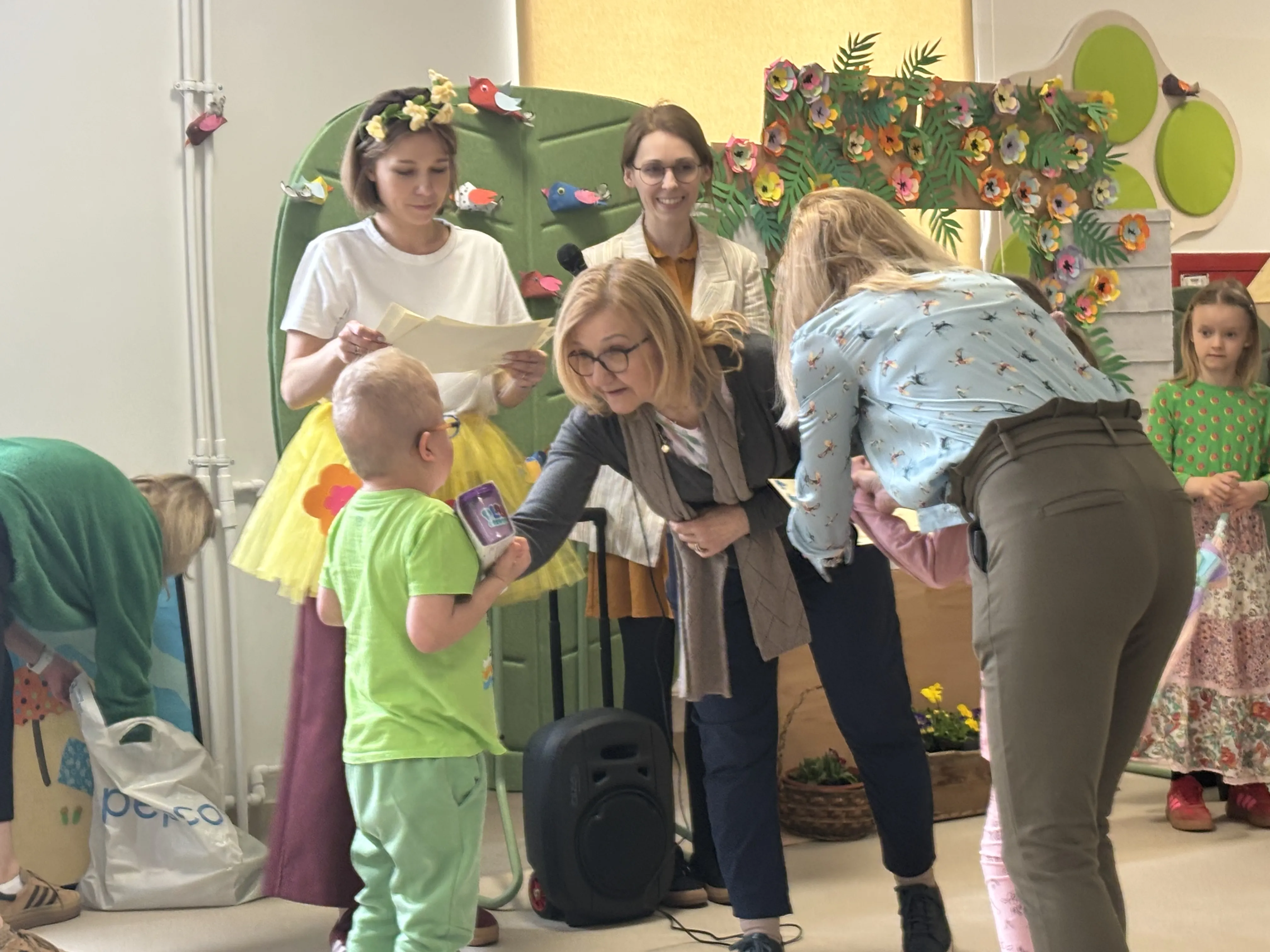 Pani wicedyrektor wręcza uczniowi nagrodę, w tle widoczna jest druga pani wicedyrektor oraz nauczycielka,  która trzyma w ręku dyplomy. Za paniami widoczne są kolorowe dekoracje