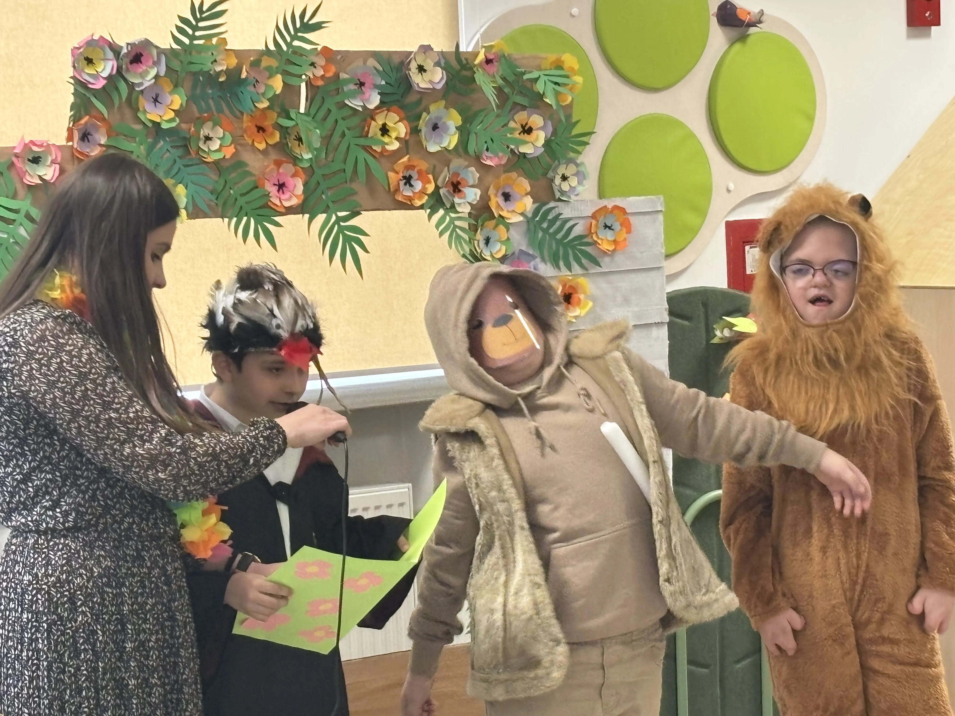 Występ uczniów: przy mikrofonie stoi elegancko ubrany uczeń, obok pani nauczycielka, która trzyma mu przy ustach mikrofon. Uczeń trzyma w ręku rozłożoną kolorową kartę, obok niego stoi dwóch innych uczniów w przebraniach.  Ponad ich głowami widoczne są kolorowe kwiaty.