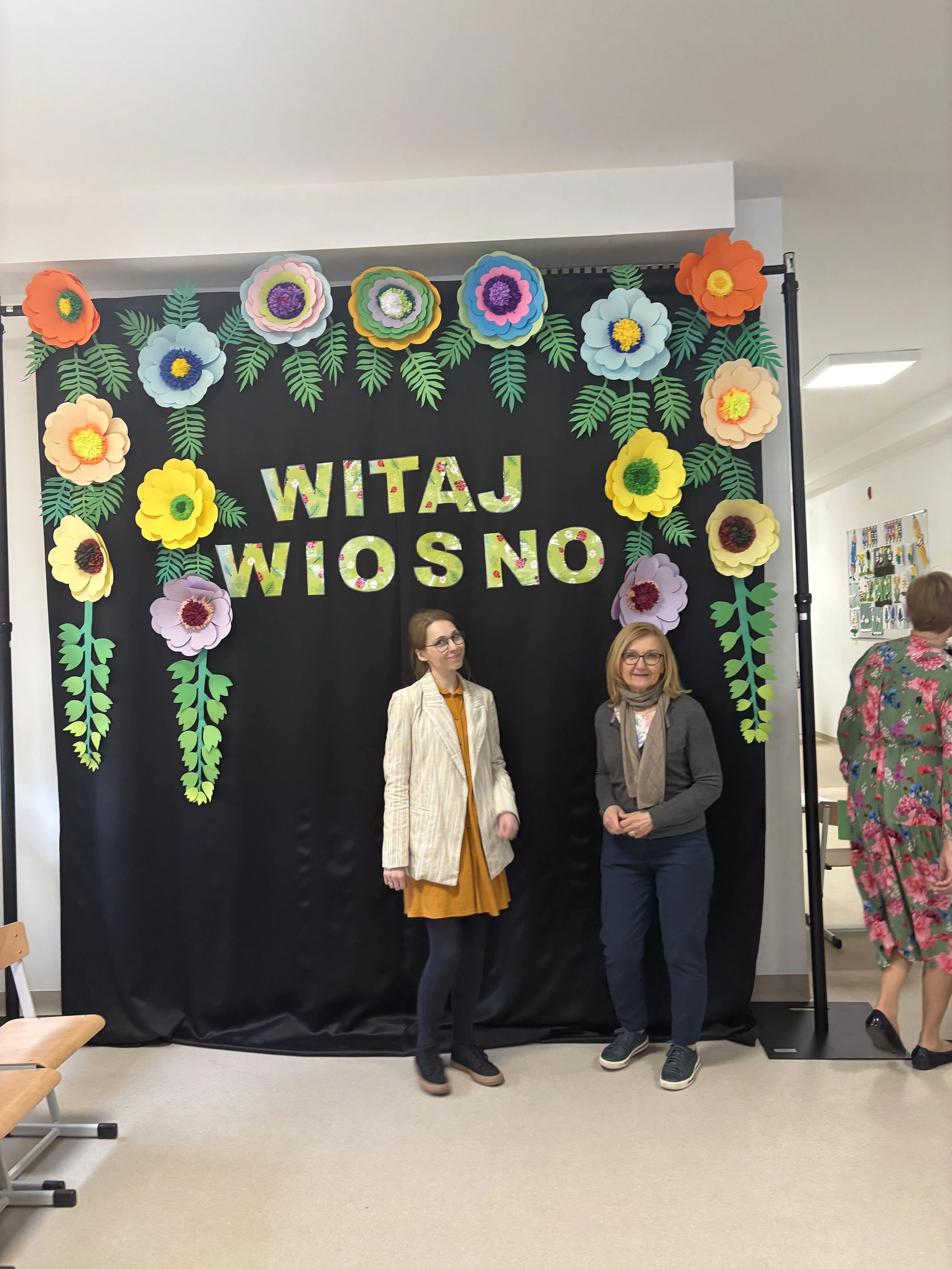 Panie wicedyrektorki szkoły podst.  nr 147 stoją na korytarzu szkolnym na tle wiosennej dekoracji z napisem Witaj Wiosno 