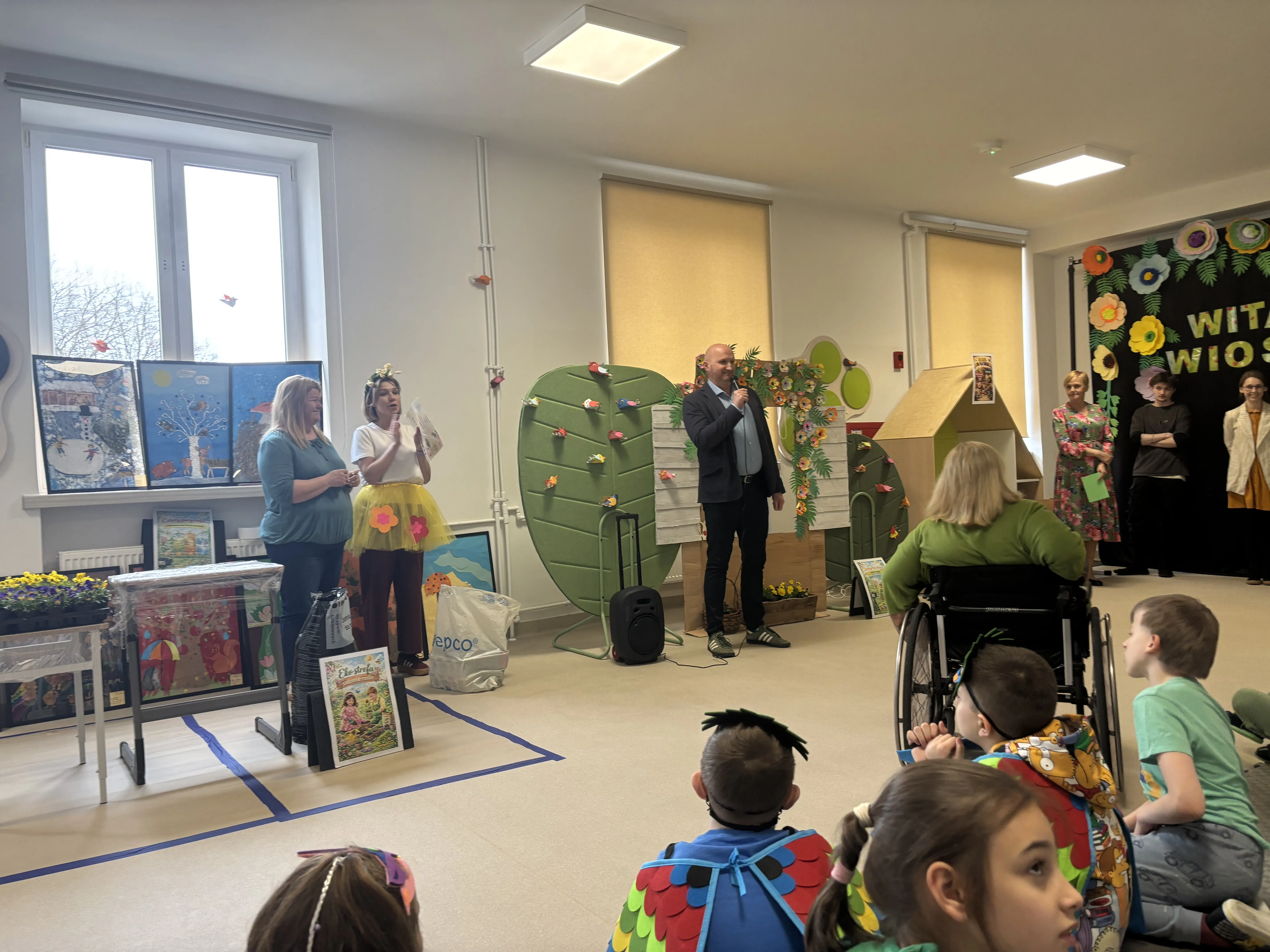 Dyrektor szkoły stoi  na korytarzu szkolnym przed dziecięcą publicznością. W tle widoczne są barwne dekoracje. Trochę dalej stoją dwie panie nauczycielki. Przy nich widoczne są kolorowe obrazy oraz skrzynki z kwiatami. 