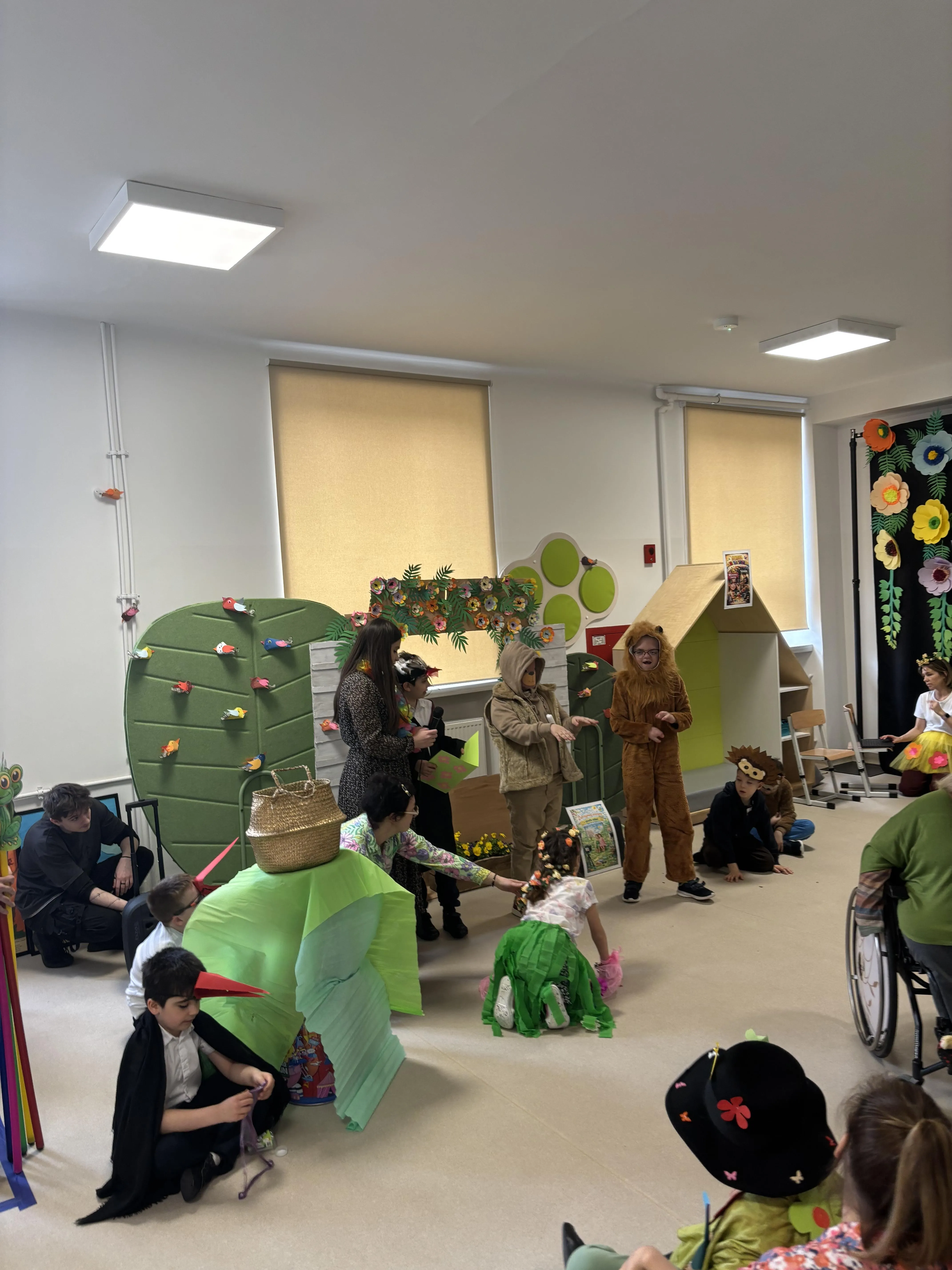 Na "scenie" znajduje się kilkoro uczniów: troje stoi  inni klęczą. Obok widoczne są  panie:  nauczycielka oraz pomoc. Jeden z klęczących uczniów ma  założony dziób bociana. Na scenie widoczne są różne rekwizyty.