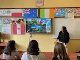 Na ścianie w klasie wisi tablica multimedialna, obok stoi studentka . W dolnej części zdjęcia widoczni są  uczniowie obserwujący tablicę. Na ścianie w klasie widoczne są rozwieszone  kartony z flagami różnych państw.
