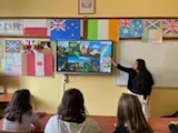 Na ścianie w klasie wisi tablica multimedialna, obok stoi studentka , która wskazuje coś na tablicy. W dolnej części zdjęcia widoczni są  uczniowie obserwujący tablicę. Na ścianie w klasie widoczne są rozwieszone  kartony z flagami różnych państw.