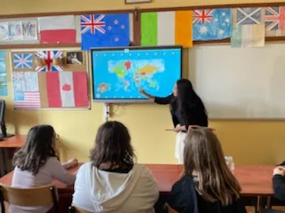 Na ścianie w klasie wisi tablica multimedialna, obok stoi studentka , która pokazuje coś na tablicy. W dolnej części zdjęcia widoczni są  uczniowie obserwujący tablicę. Na ścianie w klasie widoczne są rozwieszone  kartony z flagami różnych państw.