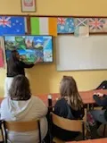 Na ścianie w klasie wisi tablica multimedialna, obok stoi studentka . W dolnej części zdjęcia widoczni są  uczniowie obserwujący tablicę. Na ścianie w klasie widoczne są rozwieszone  kartony z flagami różnych państw.