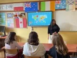 Na ścianie w klasie wisi włączona tablica multimedialna, obok stoi studentka . W dolnej części zdjęcia widoczni są  uczniowie obserwujący tablicę. Na ścianie w klasie widoczne są rozwieszone  kartony z flagami różnych państw.