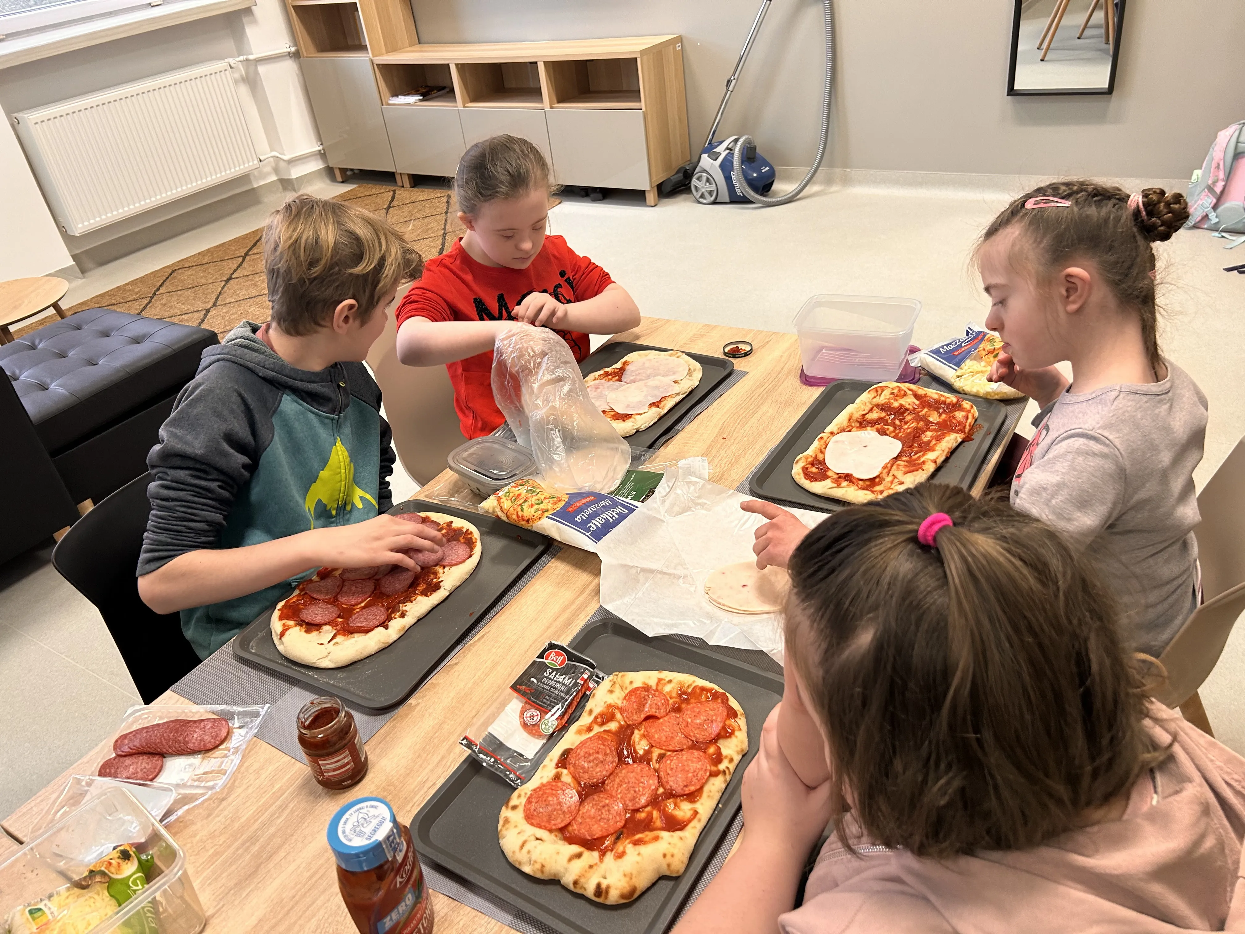 Na zdjęciu widocznych jest czterech uczniów , którzy siedzą przy stole. Przed każdym dzieckiem stoi taca, na której znajduje się upieczony  spód pizzy a  dzieci dobierają sobie dodatki : wędlinę żółty ser ketchup