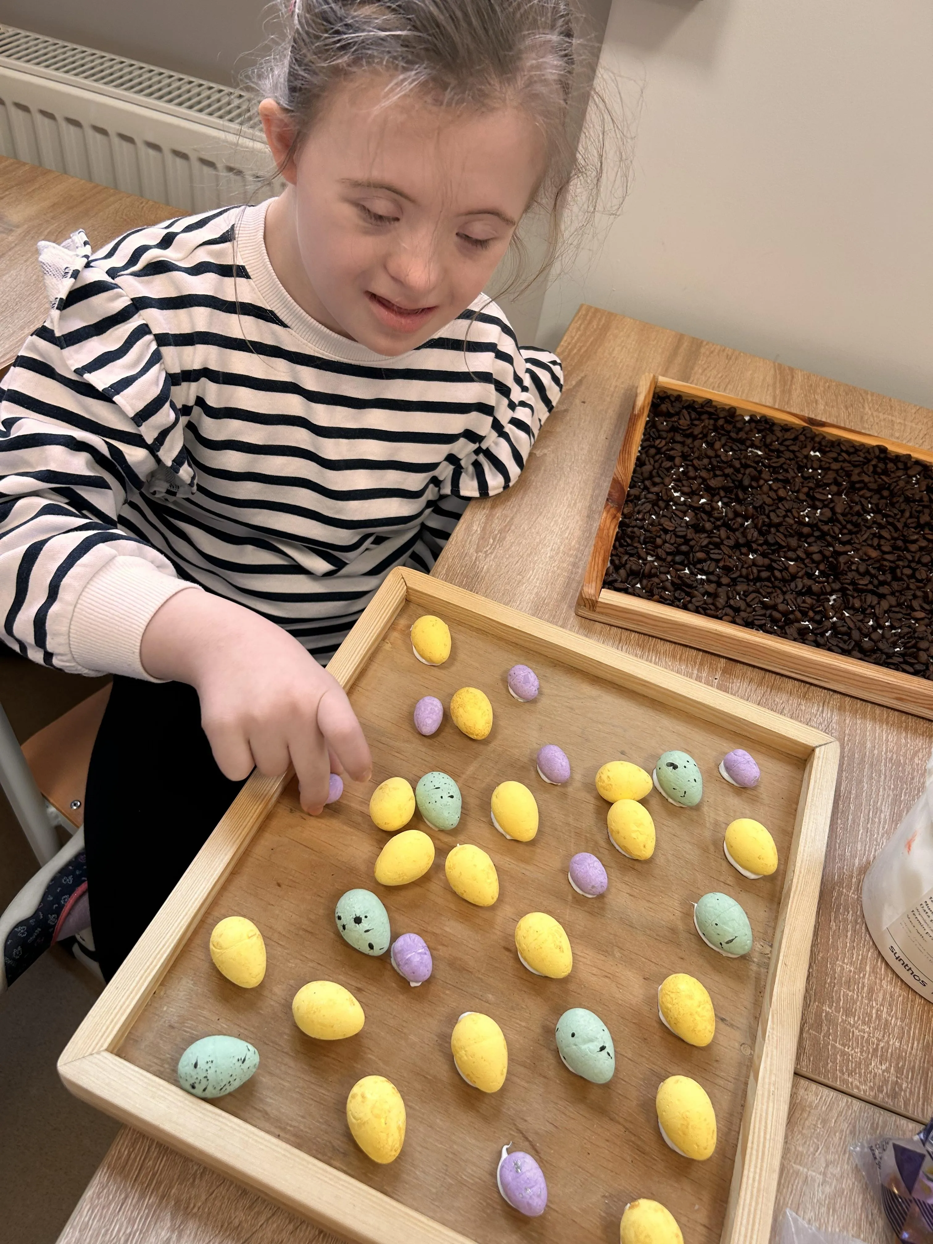 Na zdjęciu uczennica, siedząca przy drewnianej tacy, na której rozłożone są  są przedmioty w kształcie pisanek (przyklejone do podłoża) w kolorach: żółtym, fioletowym , jasnozielonym.