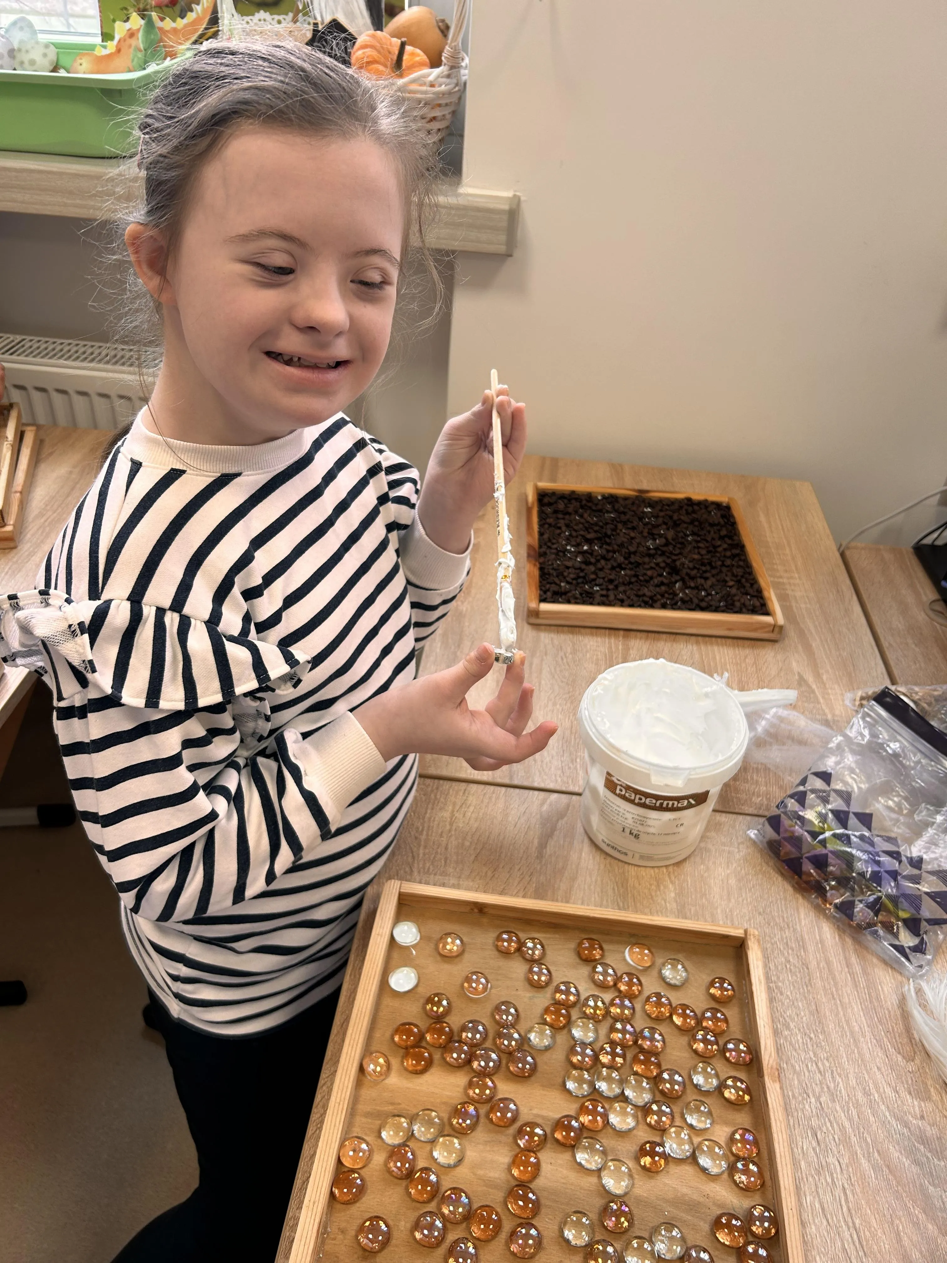 Na zdjęciu uczennica siedząca przy stoliku, na którym widać drewniane tace (w jednej znajdują sie ziarna kawy, w drugiej koralik). Na stoliku stoi również klej oraz torebka z małymi elementami . Uczennica  trzyma w ręku patyk częściowo oklejony papierem.