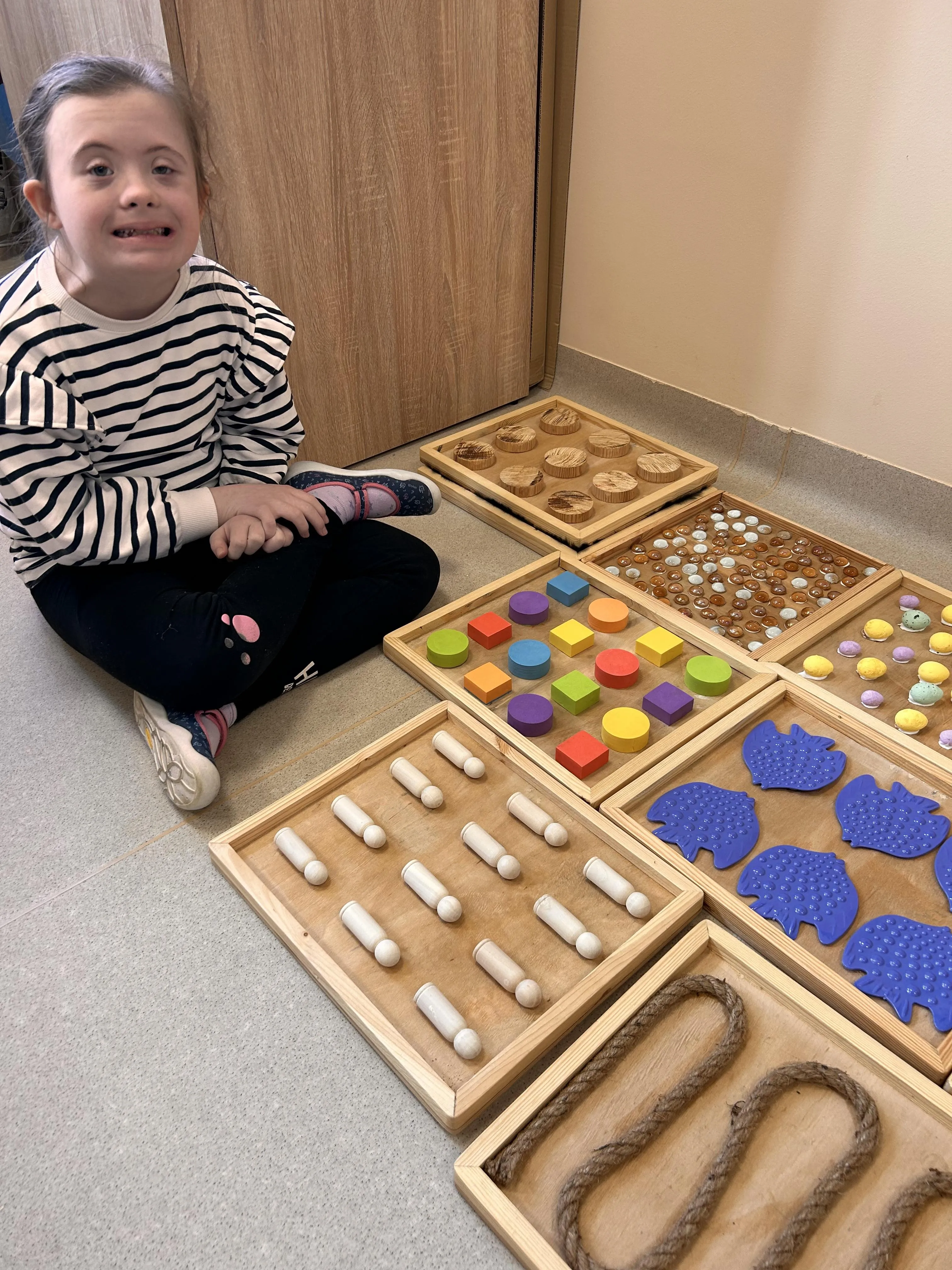 Na zdjęciu uczennica, siedząca w sali klasowej na podłodze, na której znajdują się drewniane tace . W tacach tych widoczne są przylepione do podłoża różne elementy - niektóre to kolorowe klocki  w różnych kształtach,  korale, pisanki, rybki, sznur.  