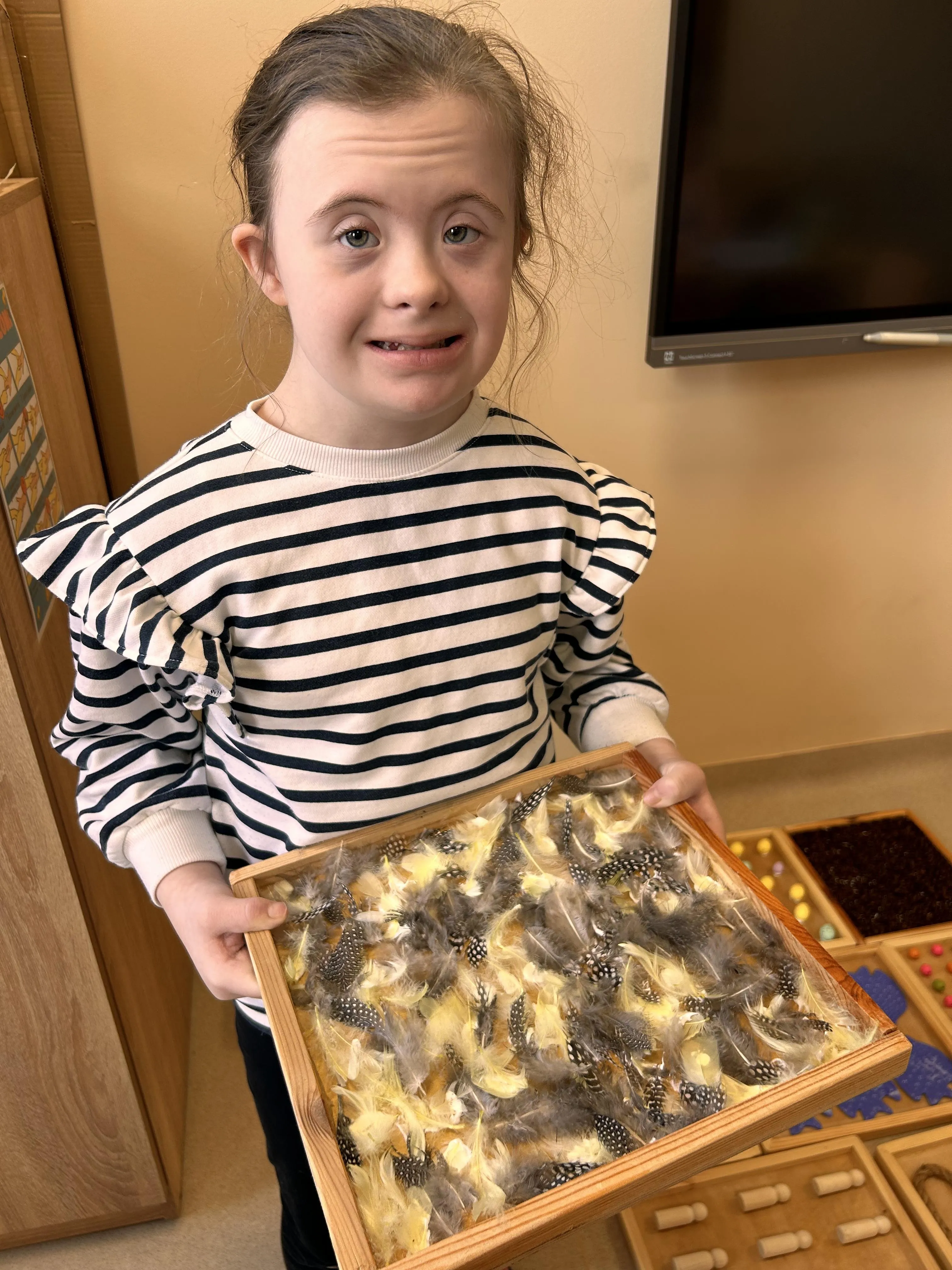 Na zdjęciu uczennica, która trzyma w ręku drewnianą tacę wypełnioną  przypominającymi ptasie pióra elementami.  Pod  tacą, którą trzyma w ręku znajdują się inne tace  z innymi  elementami.