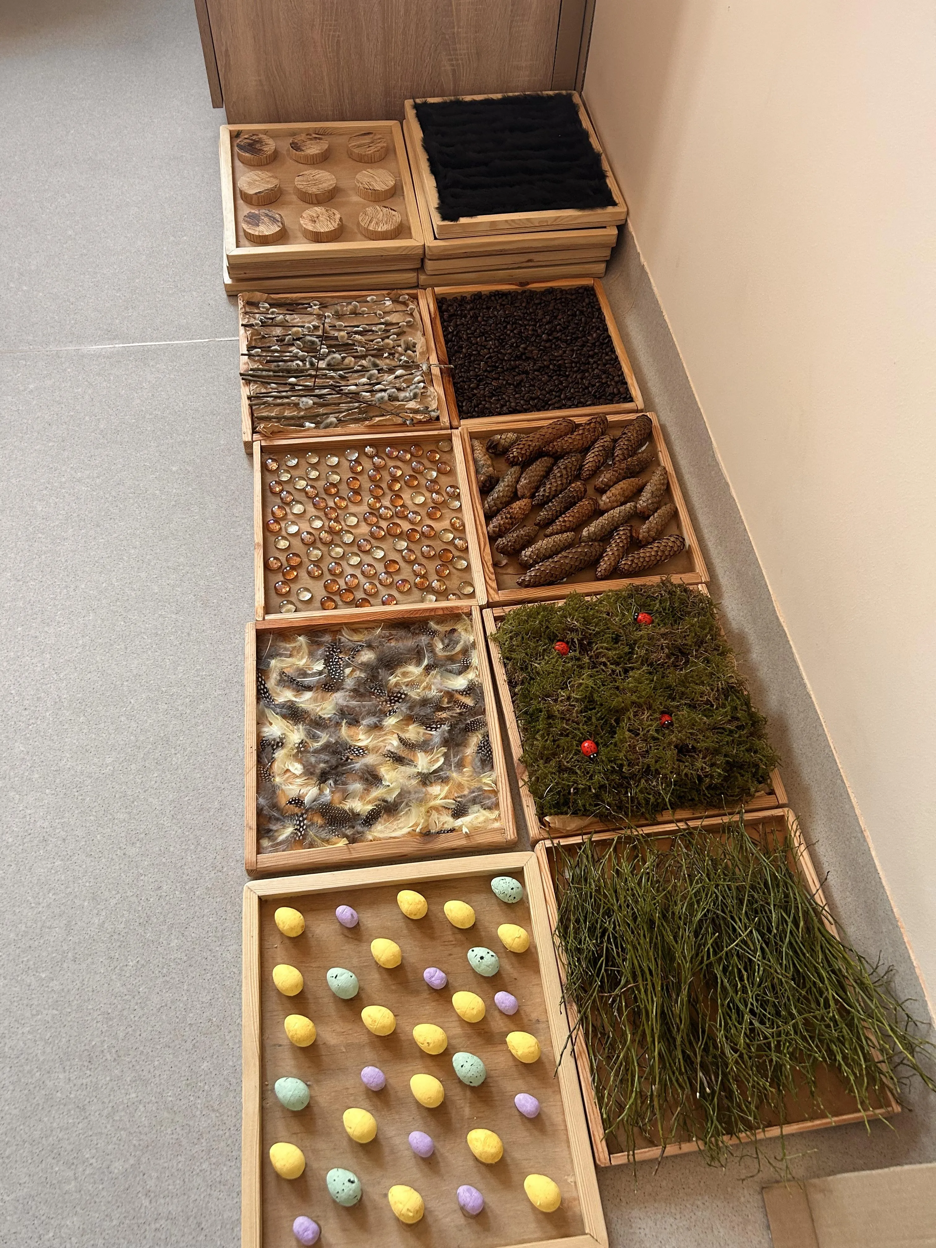 Na zdjeciu widoczne są drewniane tace , w których znajdują się  różne przedmioty: pisanki, ptasie pióra, koraliki, drewniane krążki  oraz elementy roślinne takie jak: mech, bazie, szyszki, gałązki , ziarna kawy.