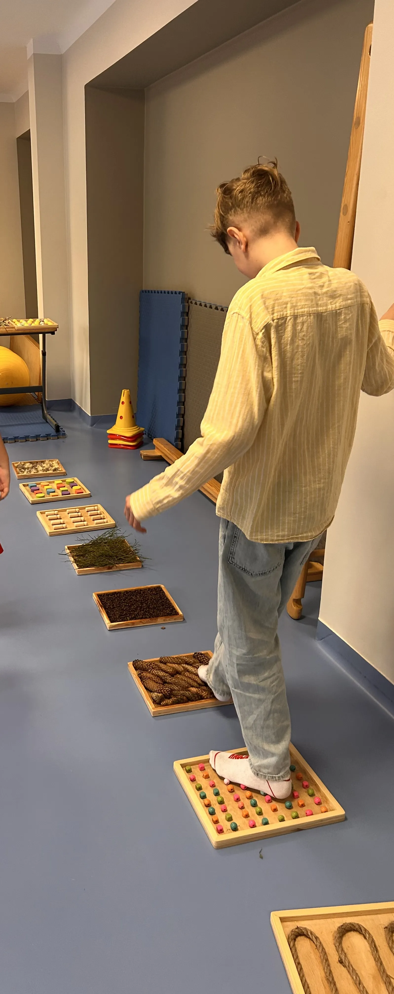 Na zdjęciu  - uczeń, który chodzi po rozłożonych na szkolnym korytarzu  drewnianych szufladach wypełnionych różnymi substancjami: szyszkami, koralikami, drewnianymi krążkami, mchem, kawą gałązkami, drewnianymi kołkami 