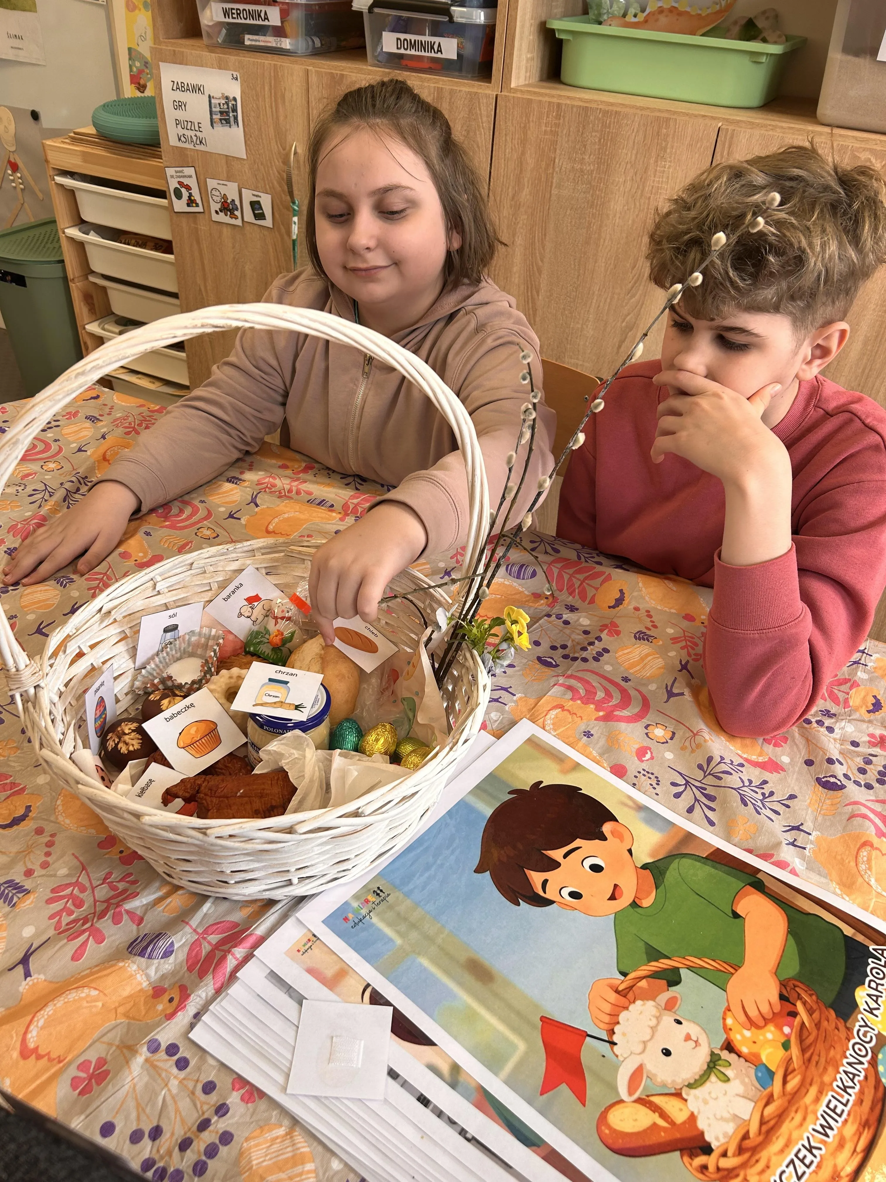 Na zdjęciu  - dwoje uczniów, którzy siedzą przy stoliku w sali lekcyjne.  Na stoliku stoi koszyczek, w którym powkładane są spożywcze produkty wielkanocne , zgodnie z karteczkami, znajdującymi się w koszyczku, przedstawiającymi  podpisane obrazki  z produktami. Koszyczek wielkanocny przyzdobiony jest baziami. Obok na stoliku leży  kartka z rysunkiem przedstawiającym chłopca, który trzyma w ręku koszyczek wielkanocny.