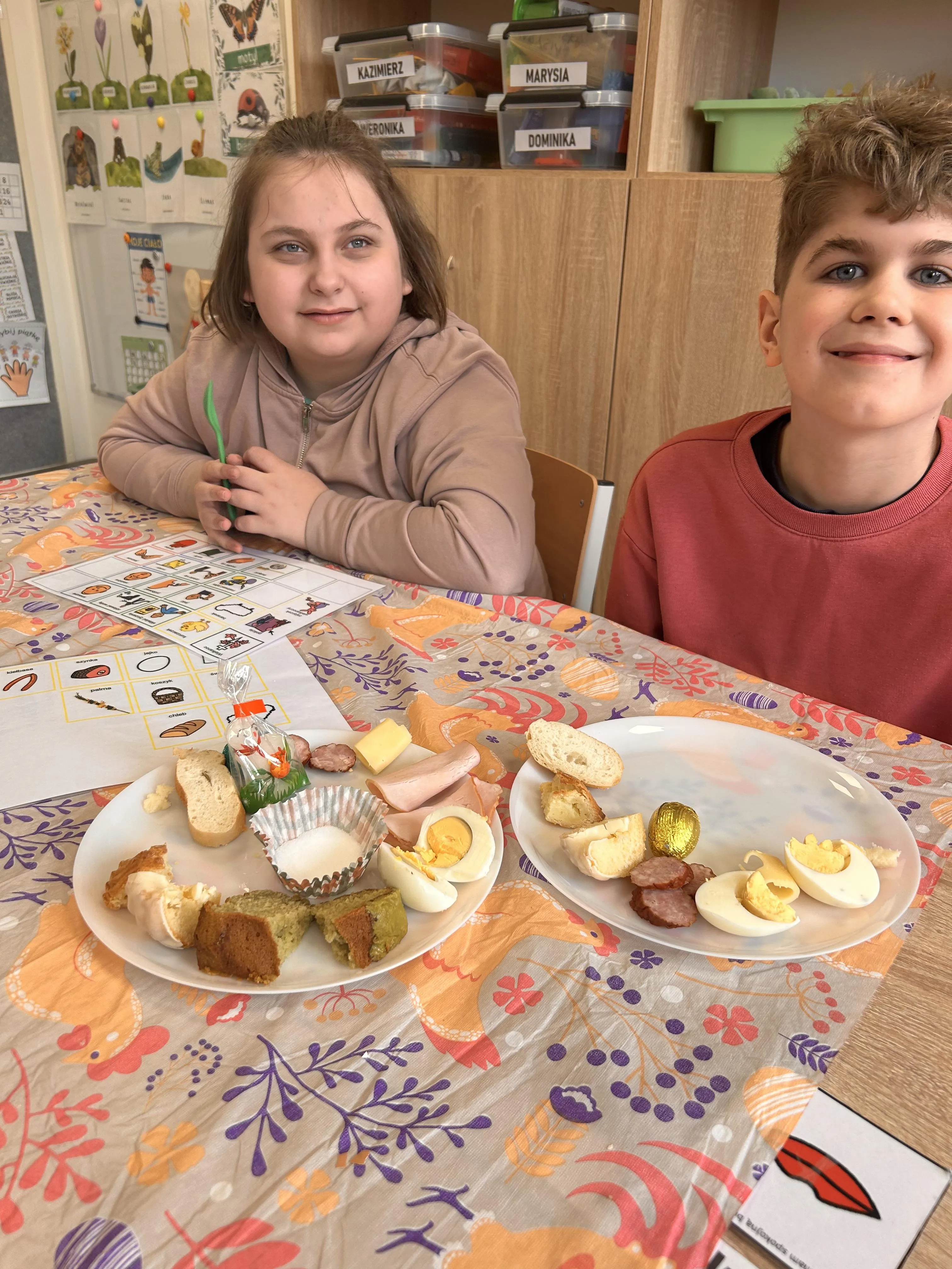 Na zdjęciu uczeń z uczennicą , siedzą przy stoliku w sali  lekcyjnej. Przed nimi stoją dwa talerzyki , na których znajdują się produkty z koszyczka  wielkanocnego.