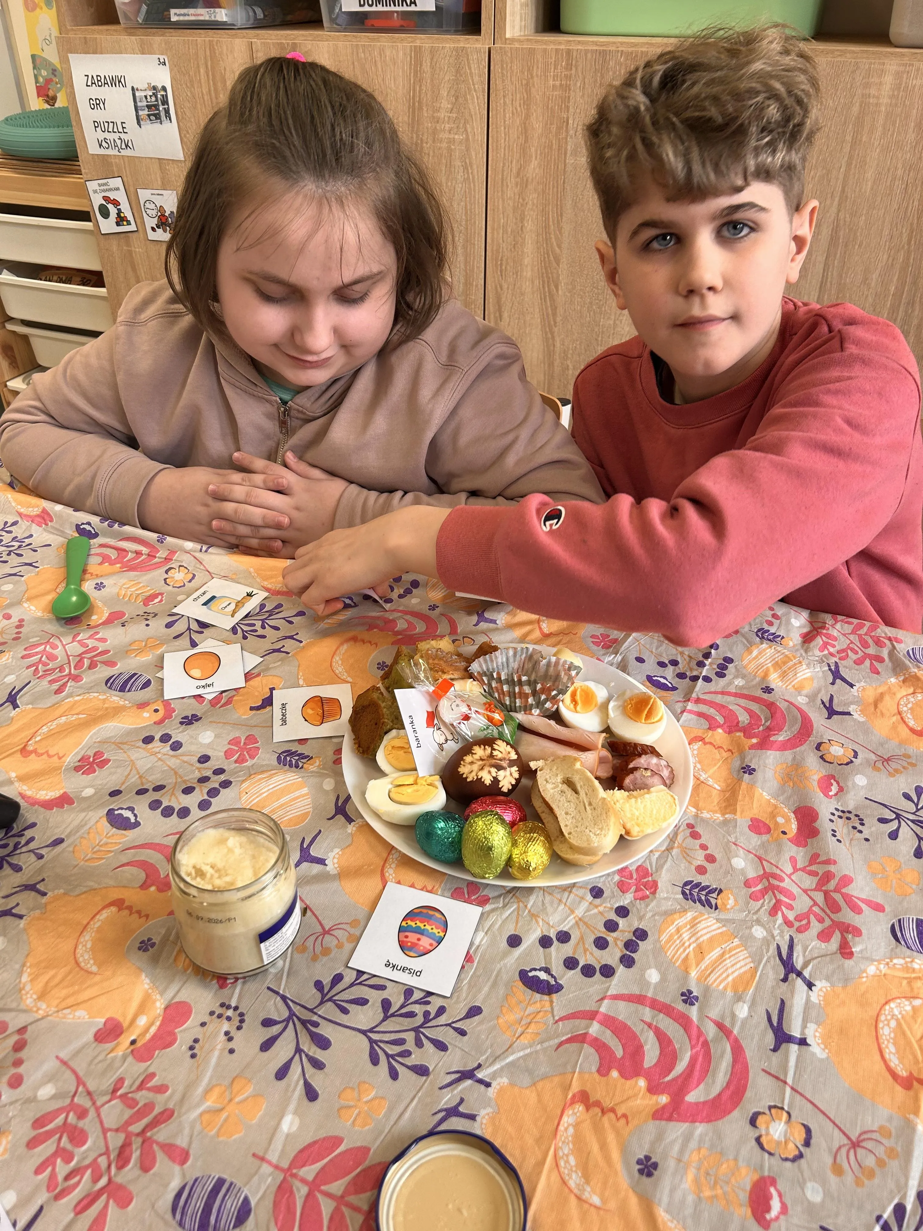 Na zdjęciu  - uczeń i uczennica,  siedzący przy stoliku na którym stoi talerzyk z produktami z koszyczka wielkanocnego. przy talerzyku stoi słoiczek z chrzanem.  Obok widoczne są  obrazkami z podpisanymi  produktami  wielkanocnymi.