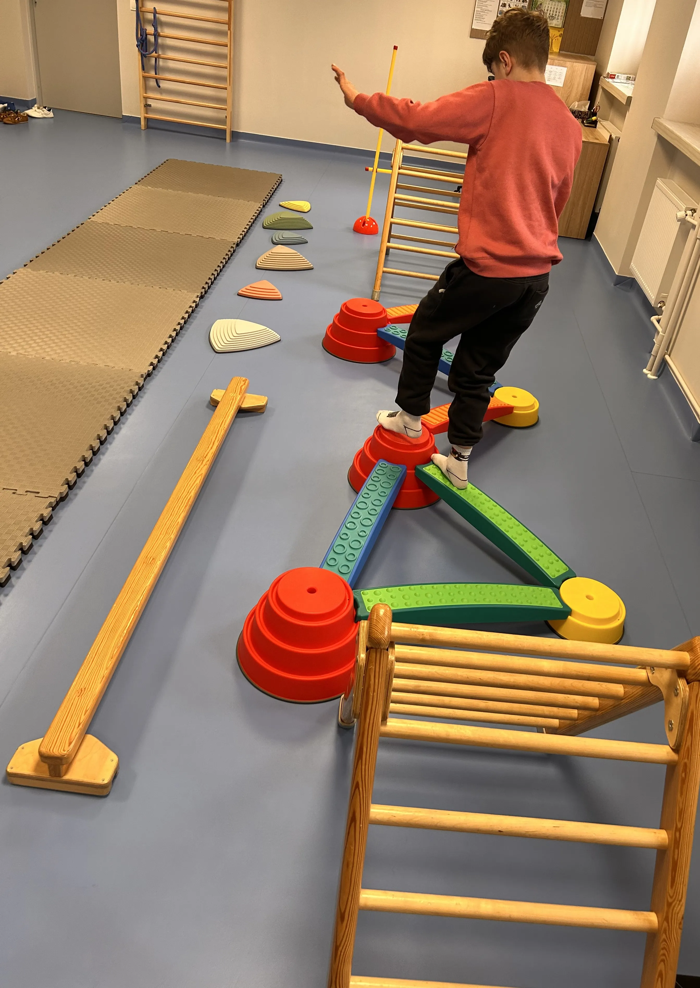 Na korytarzu szkolnym leżą  różne przedmioty . Widoczne też są stojące drabinki. Uczeń z gumowym kółkiem na głowie  stoi  na jednym z przedmiotów ,  starając się zachować równowagę.  