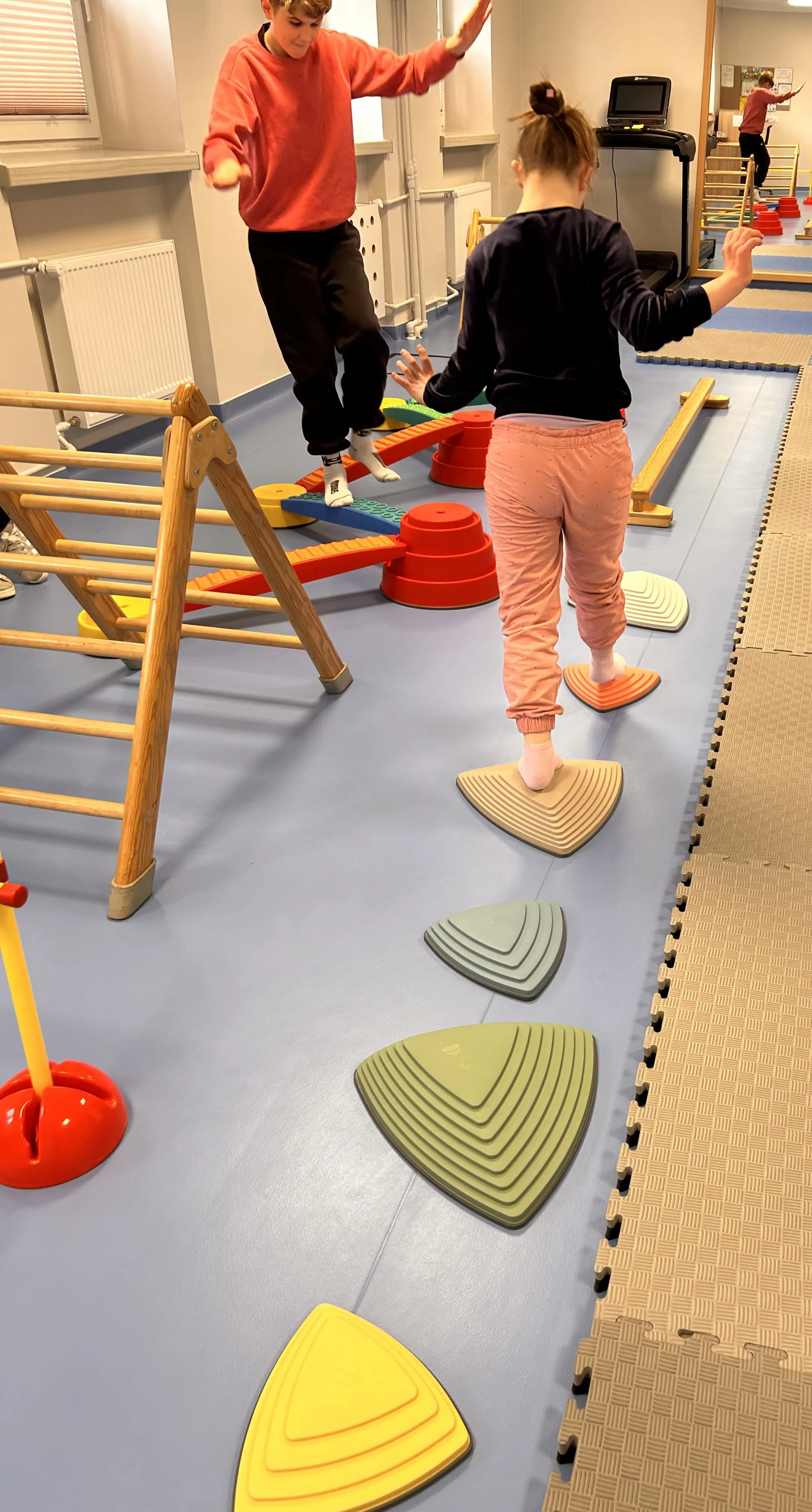 Na korytarzu szkolnym leżą  różne przedmioty , widoczna jest też stojąca drabinka.  Na zdjęciu widoczny jest  uczeń, który   stoi  na jednym z przedmiotów ,  starając się zachować równowagę. Z drugiej strony  zdjęcia widać uczennicę, która idzie po rozłożonych  gumowych przedmiotach. 