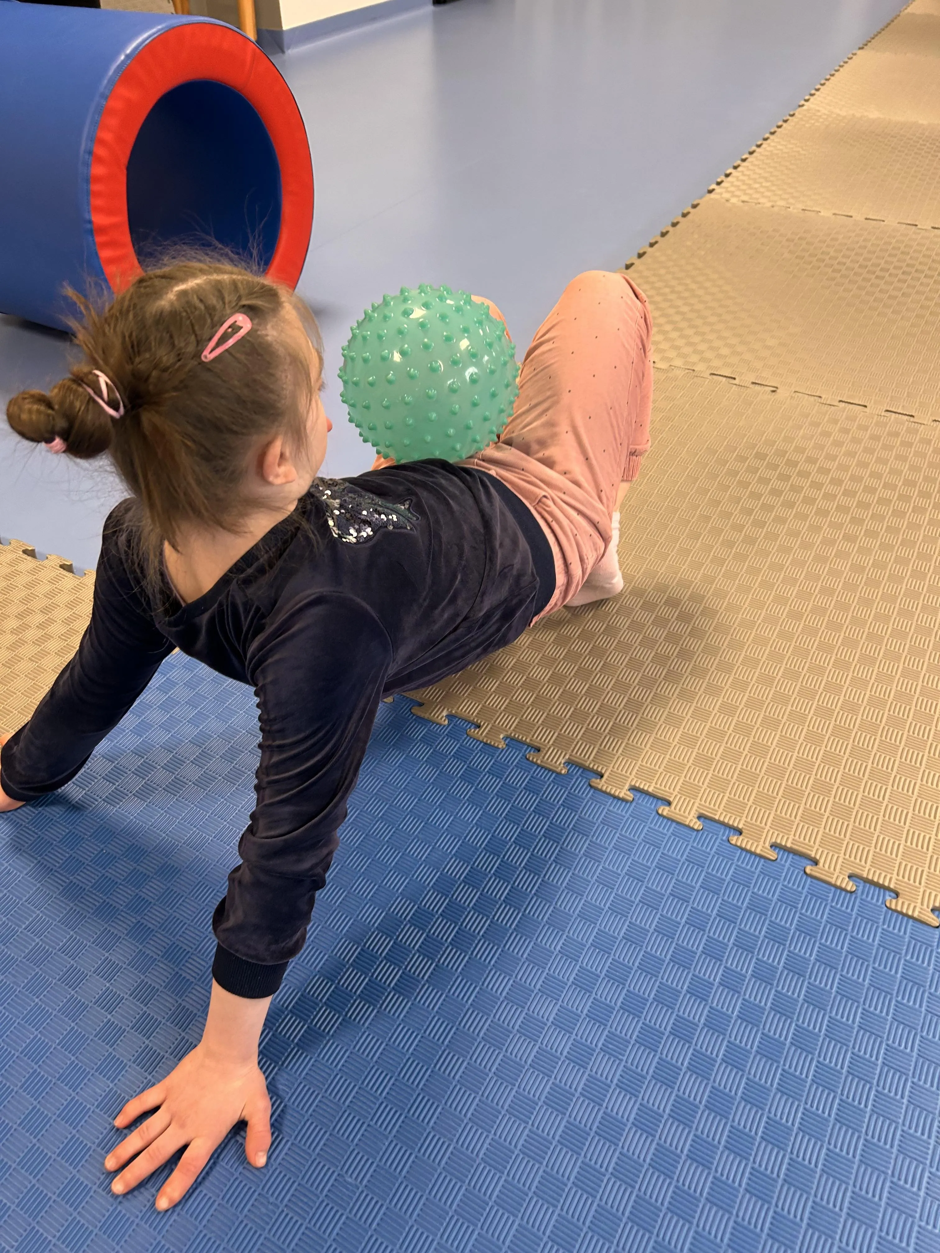 Na zdjęciu widoczna jest  uczennica (na korytarzu szkolnym), która  opiera wyciągnięte ręce i zgięte w kolanach nogi o podłogę.  Próbuje utrzymać równowagę z  leżącą na jej brzuchu piłeczką.. 