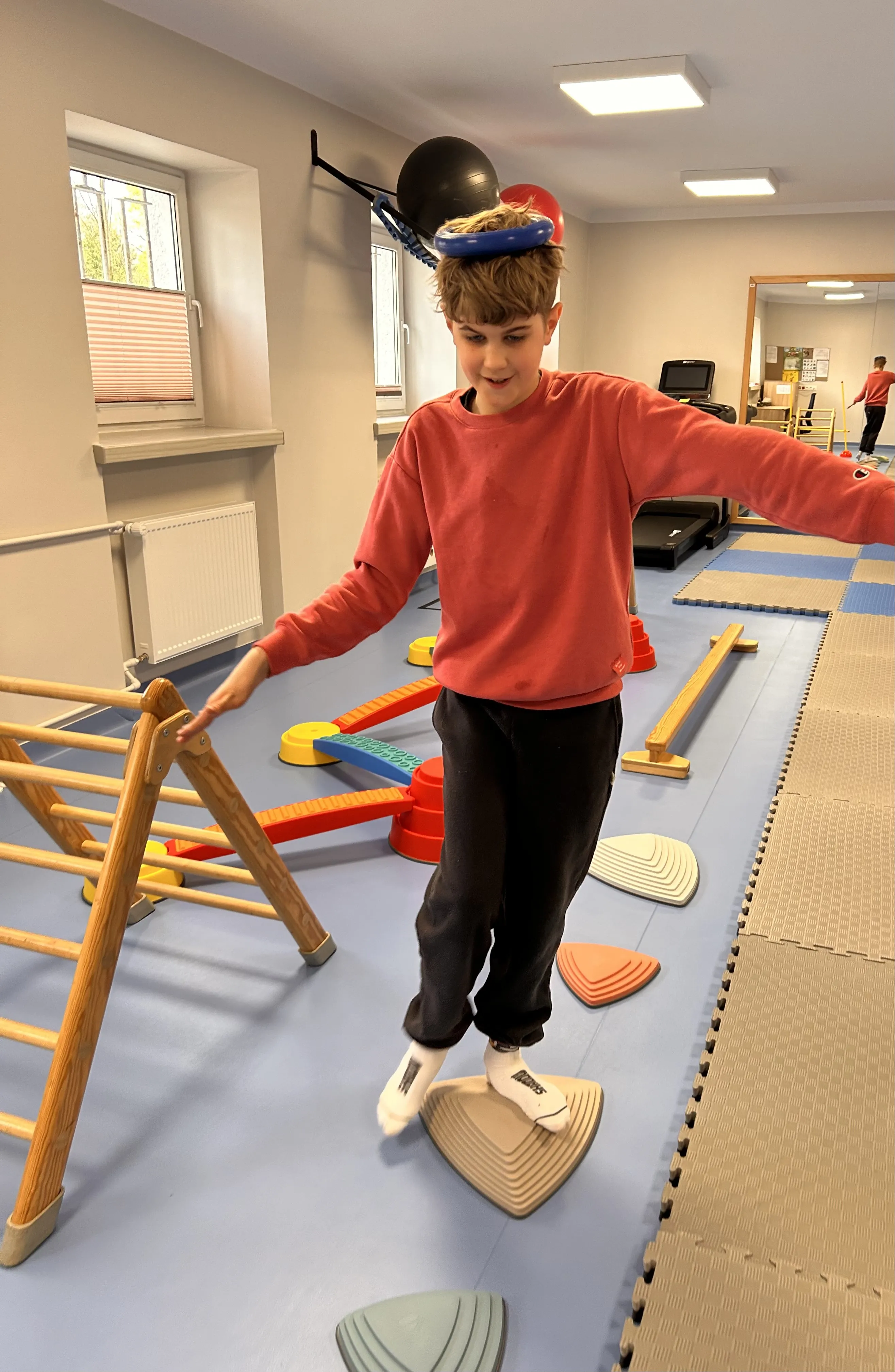 Na korytarzu szkolnym leżą  różne przedmioty , widoczna jest też stojąca drabinka.  Uczeń z gumowym kółkiem na głowie  stoi  na jednym z przedmiotów ,  starając się zachować równowagę.  