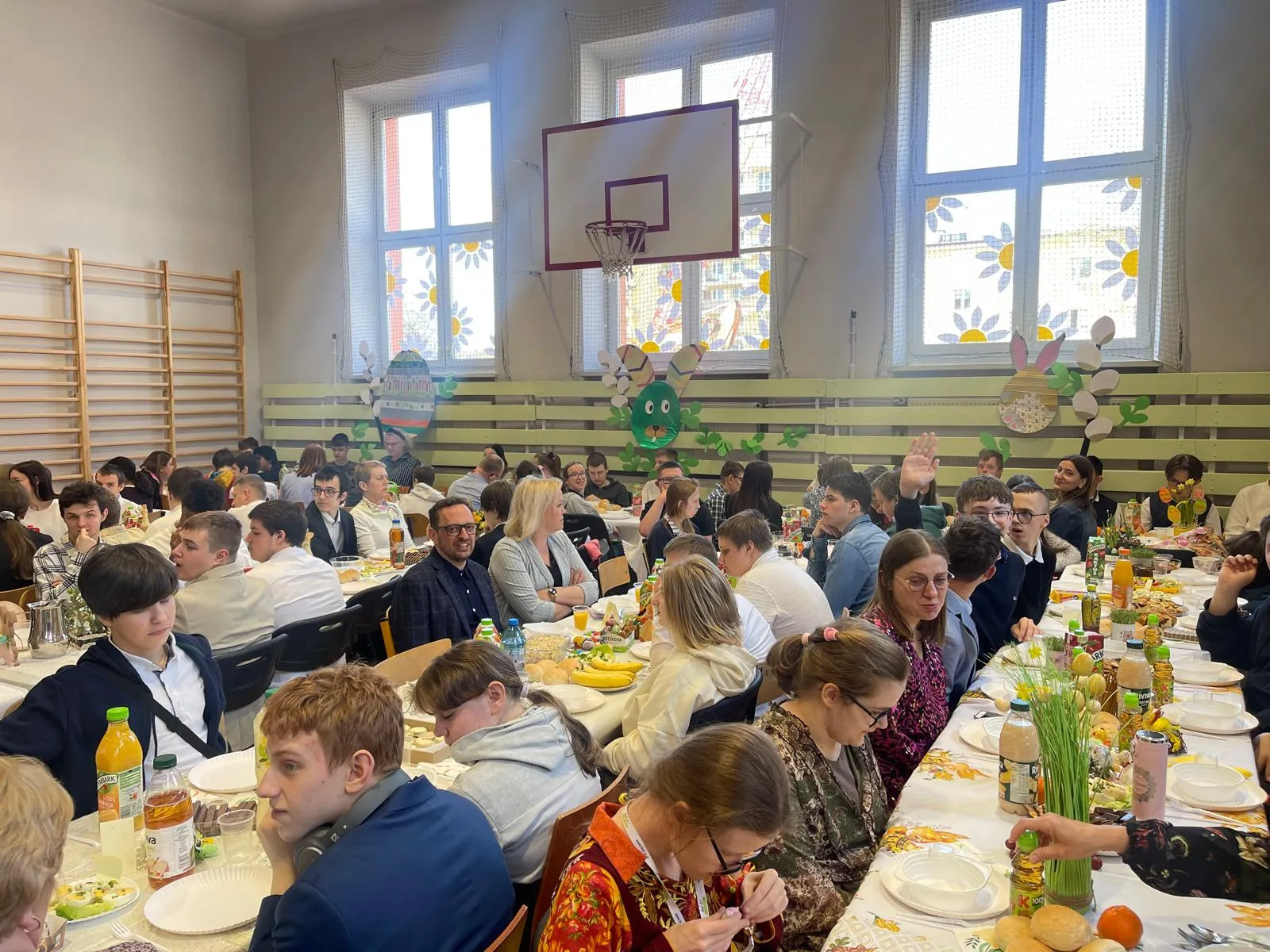 uczniowie i nauczyciele siedzacy przy stołach w sali gimnastyczne przy śniadaniu wielkancnym