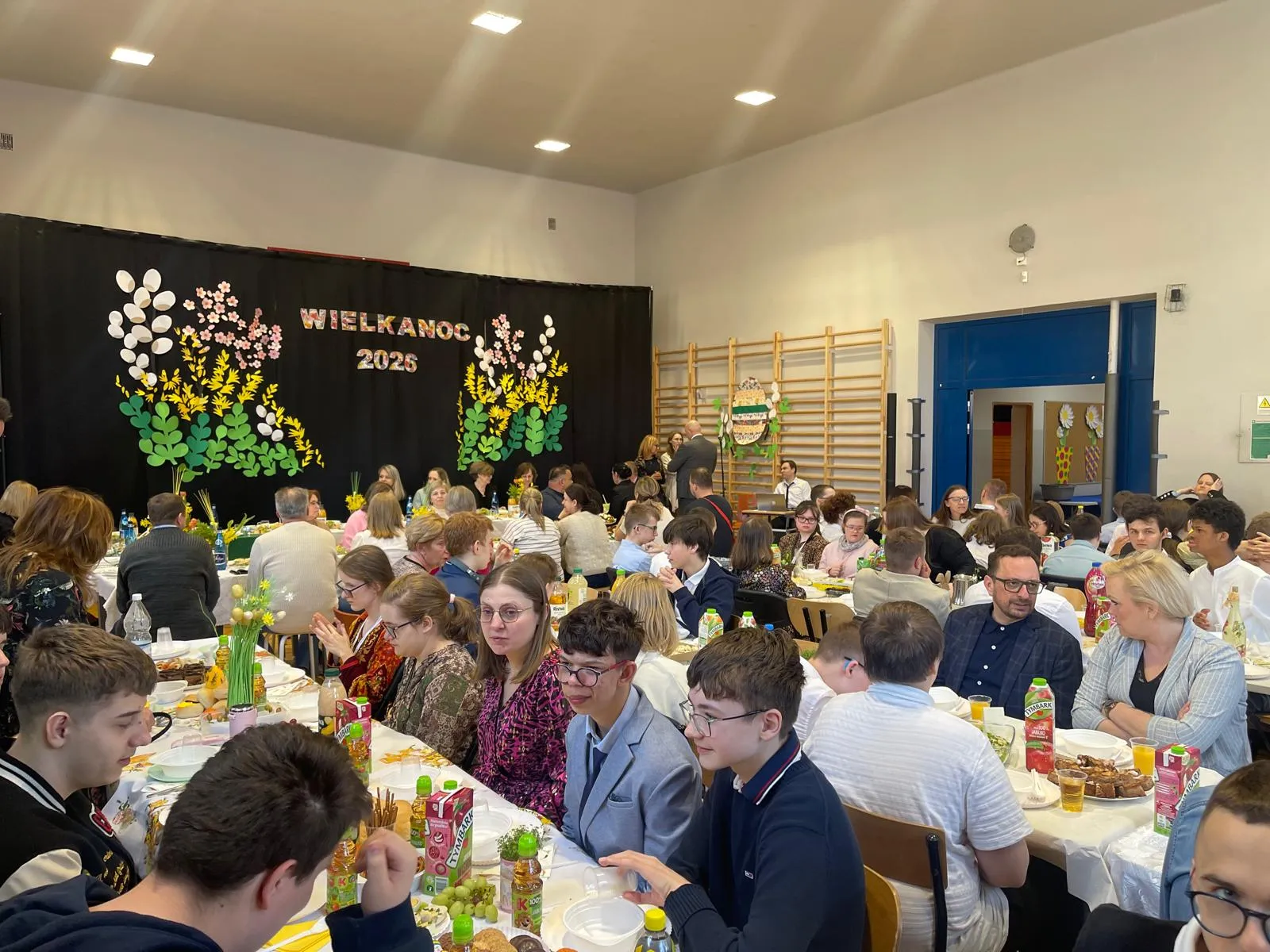 uczniowie i nauczyciele siedzacy przy stołach w sali gimnastycznej przy śniadaniu wielkancnym