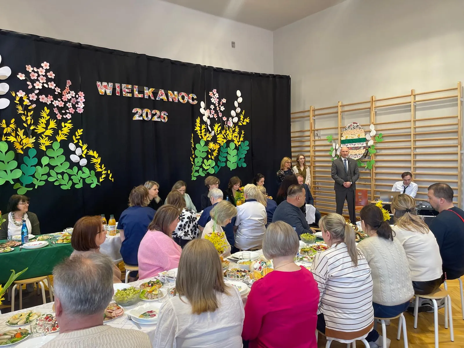 pracownicy szkoły siedzacy przy stołach w tle przemawiajacy dyrektor