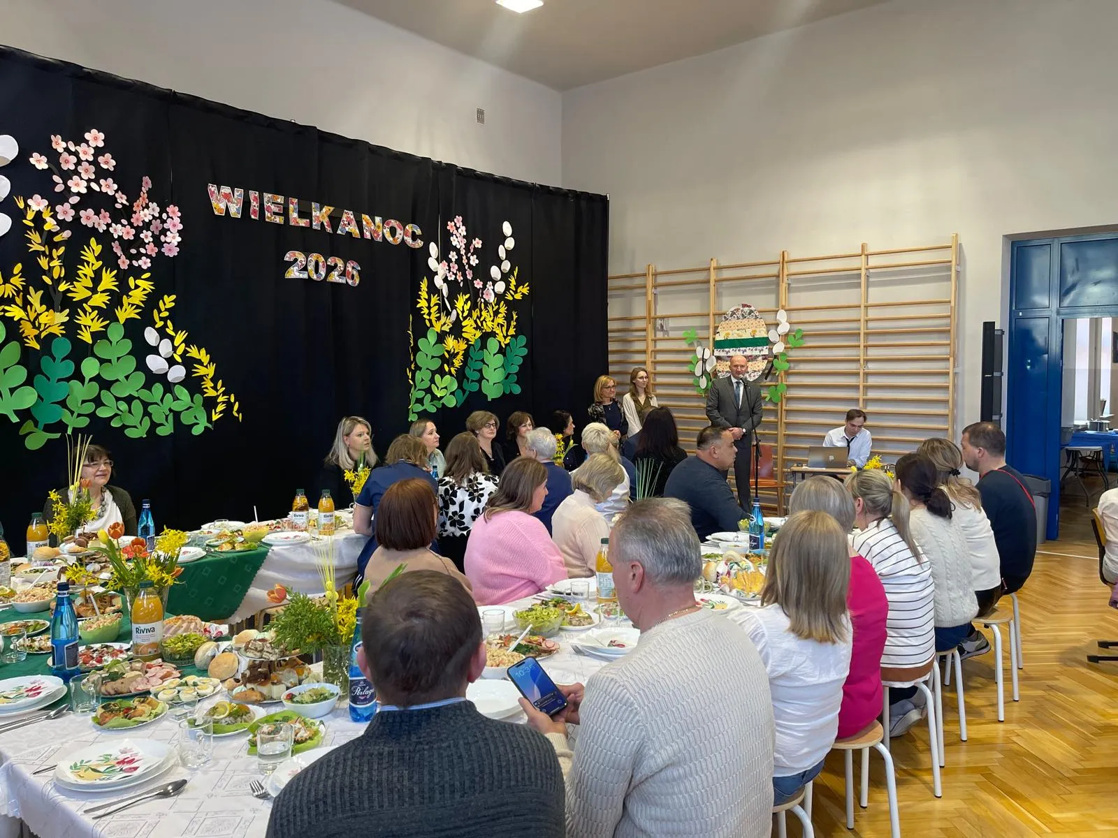 uczniowie i nauczyciele siedzacy przy stołach w sali gimnastyczne przy śniadaniu wielkancnym