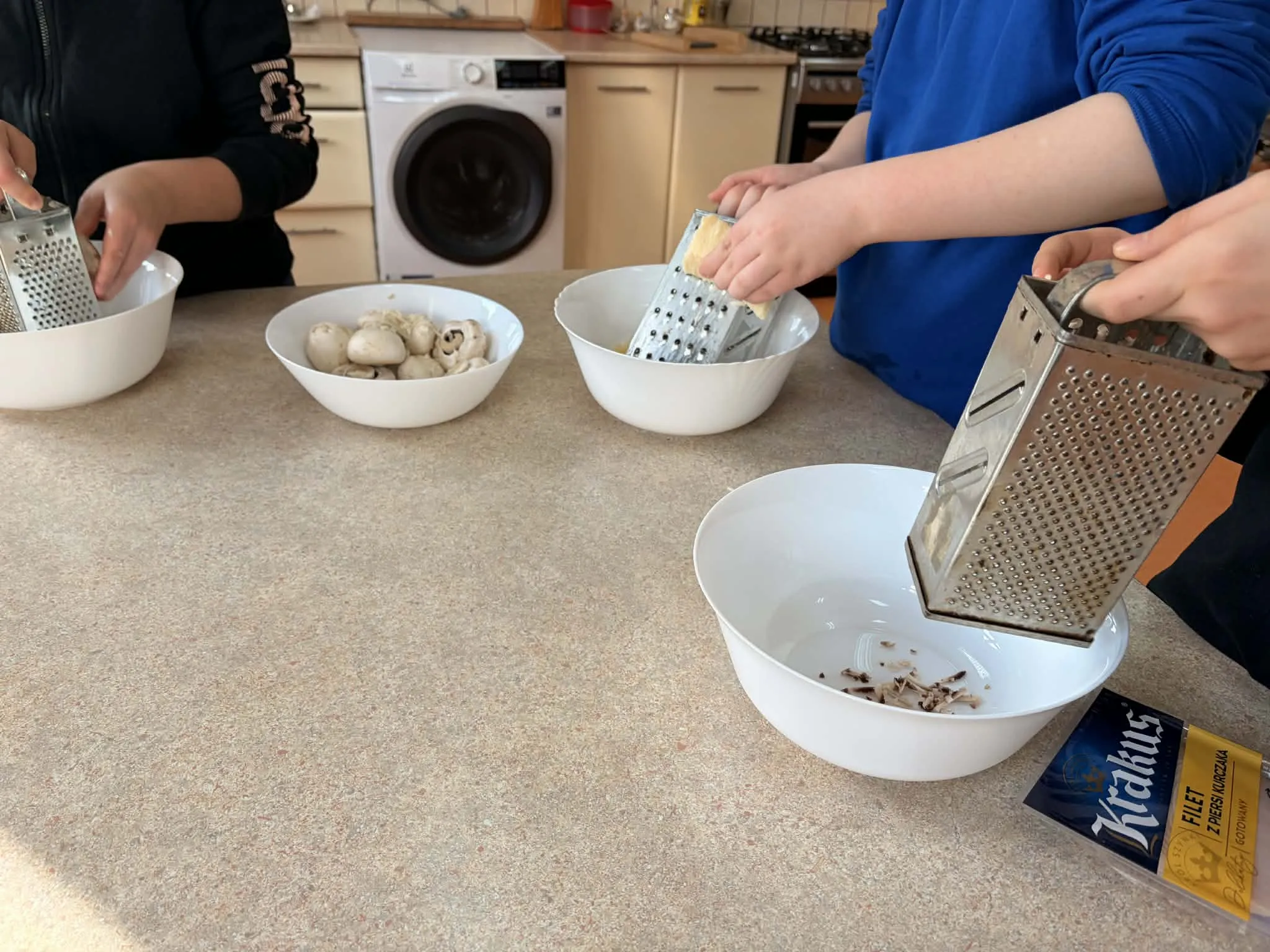 Na pierwszym planie  - stół , znajdujący się w pomieszczeniu gospodarczym. Na stole są  cztery miski. W jednej są pieczarki, a trzy pozostałe wypełniają się kawałkami pieczrek, które trą na tarkach uczniowie (na zdjęciu widoczne są  ręce uczniów.