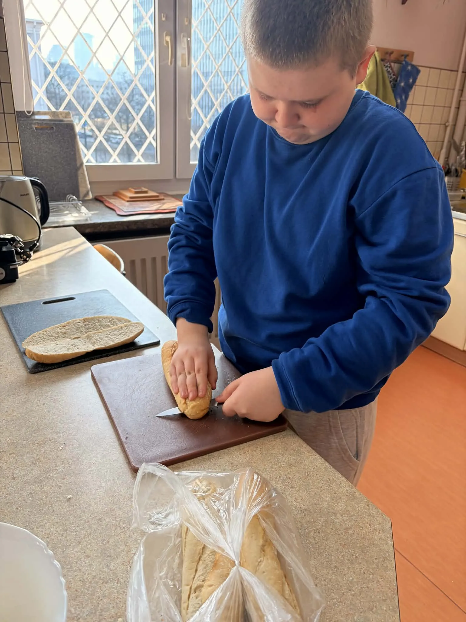 Na zdjęciu widoczny jest uczeń, stojący przy blacie  kuchennym w pomieszczeniu gospodarczym. Chłopiec na desce kroi na pół bagietkę. Na desce obok leży połówka bagietki. Przy chłopcu stoi woreczek z bagietkami.  