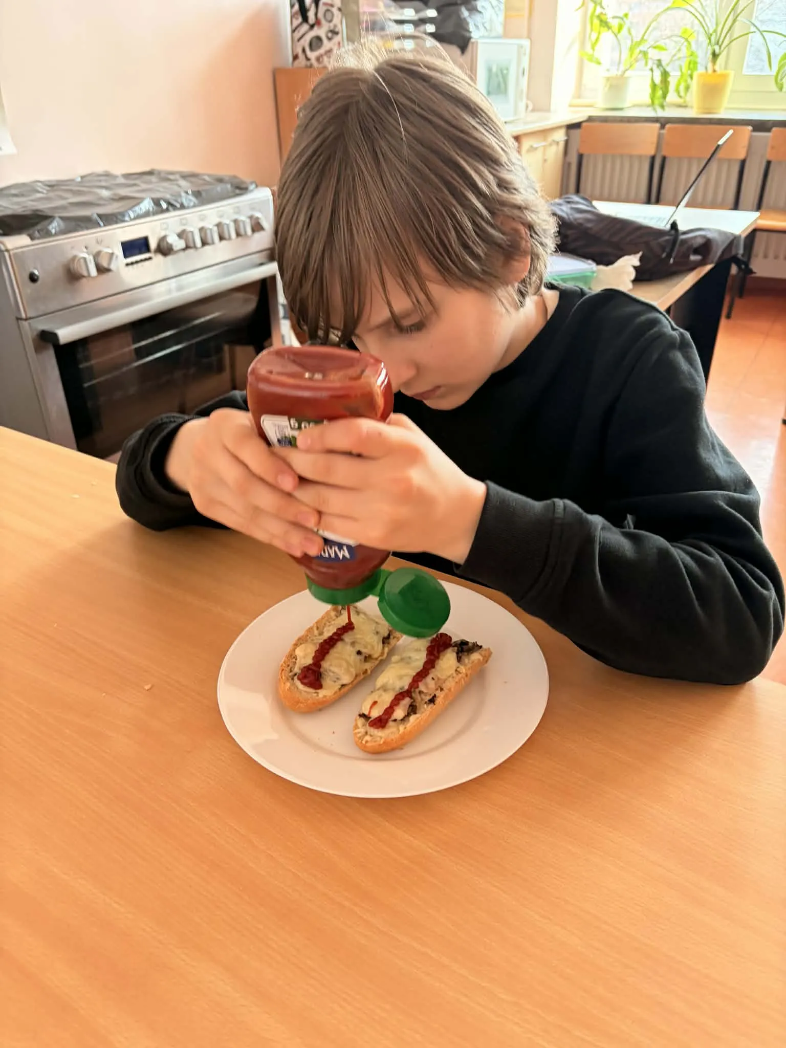 Na zdjęciu uczeń siedzący przy stole w pomieszczeniu gospodarczym. Przed chłopcem stoi talerz, na którym leży przekrojona na pół zapiekanka. Chłopiec wyciska ketchup na zapiekanki.  