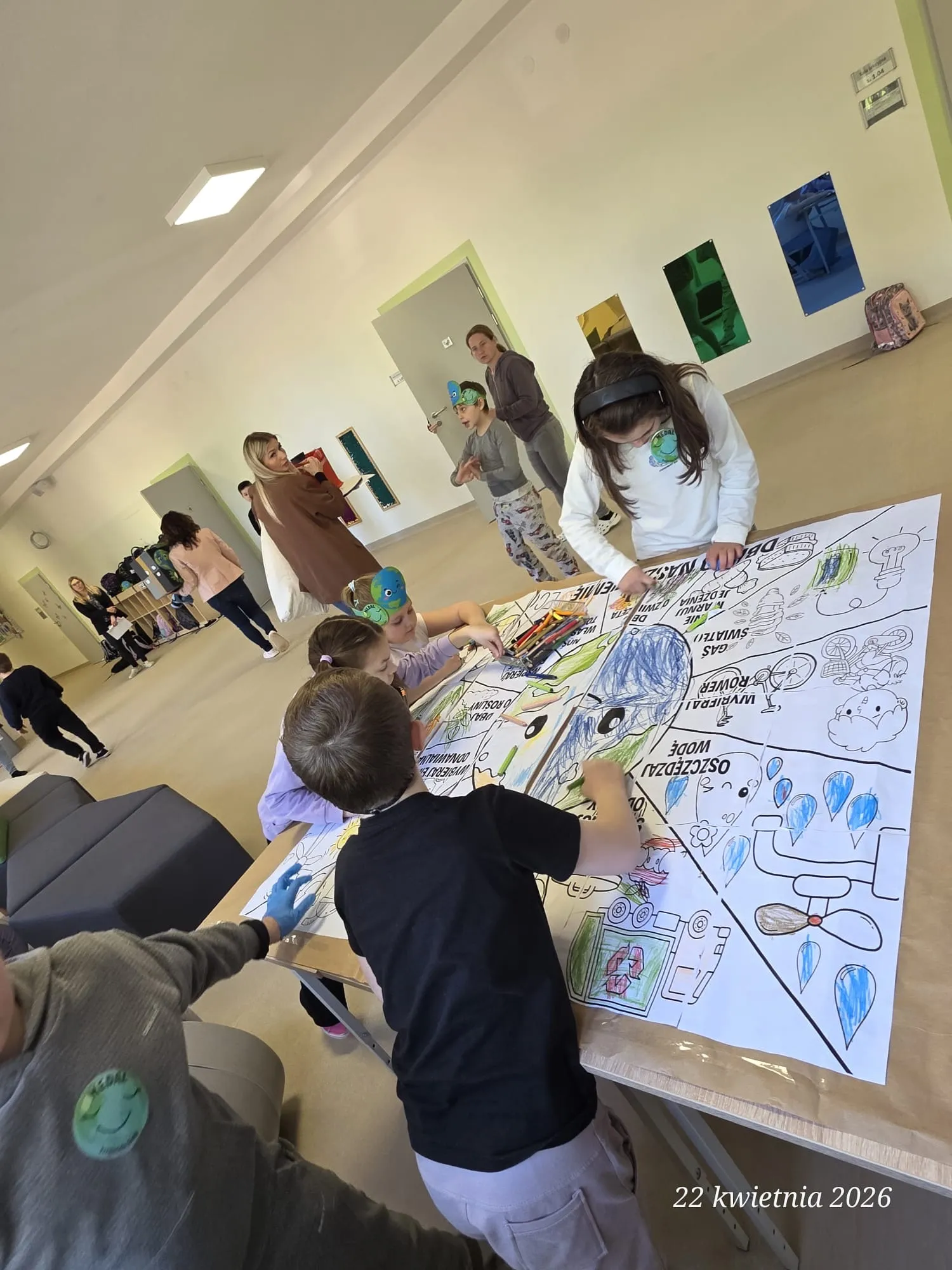 Na zdjęciu grupa uczniów pochylonych nad leżącą na stole kolorowanką. Uczniowie mają do dyspozycji kredki. W tle widoczni są inni uczniowie  i nauczyciele .