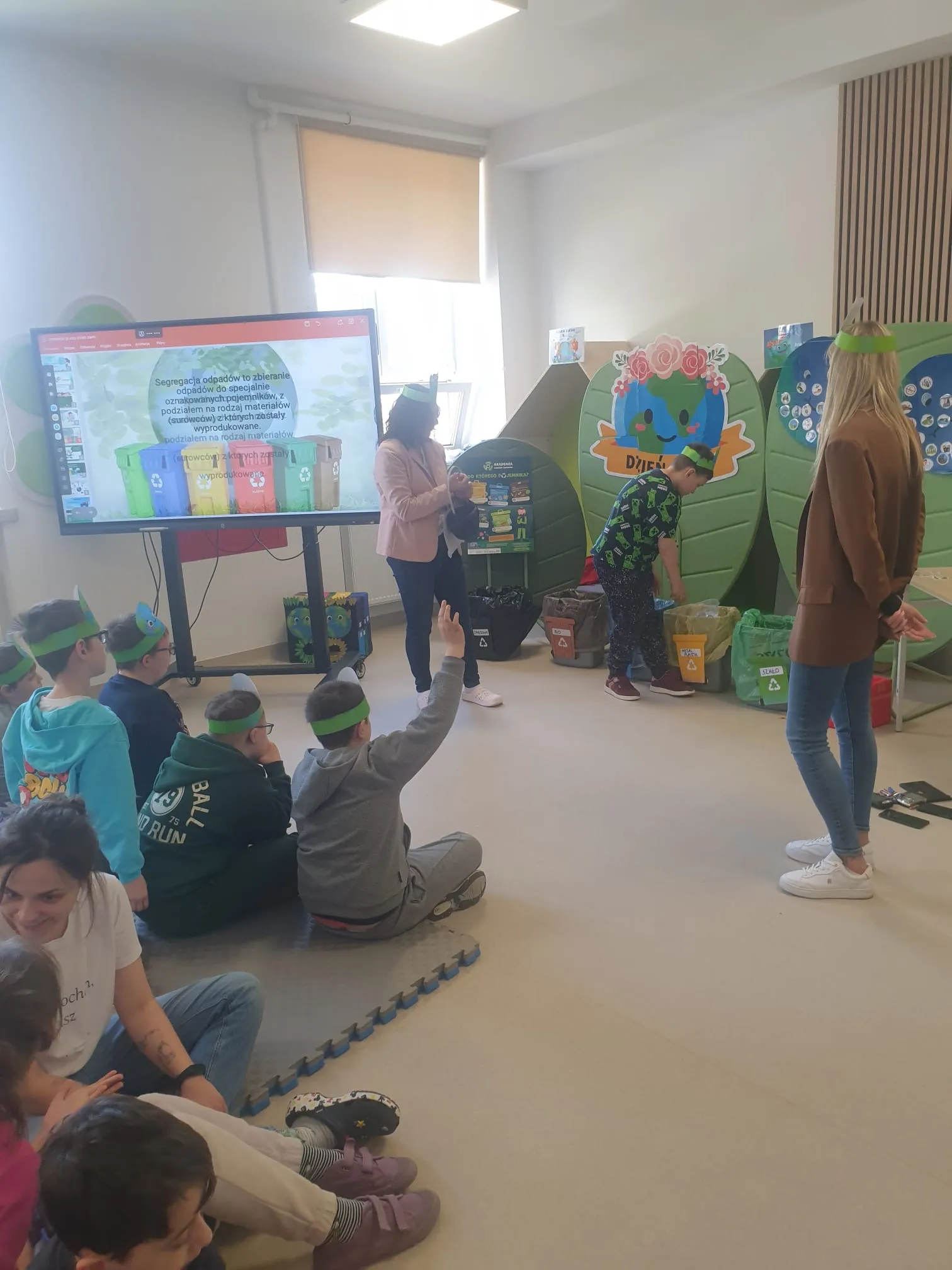 Na zdjęciu widoczni są uczniowie siedzący przed tablicą multimedialna na korytarzu szkolnym. Na tablicy widoczny jest slajd z koszami  na odpady. Kosze w tych samych kolorach ustawione są na korytarzu. Do jednego z nich uczeń coś wrzuca, w pobliżu stoją dwie panie nauczycielki.
