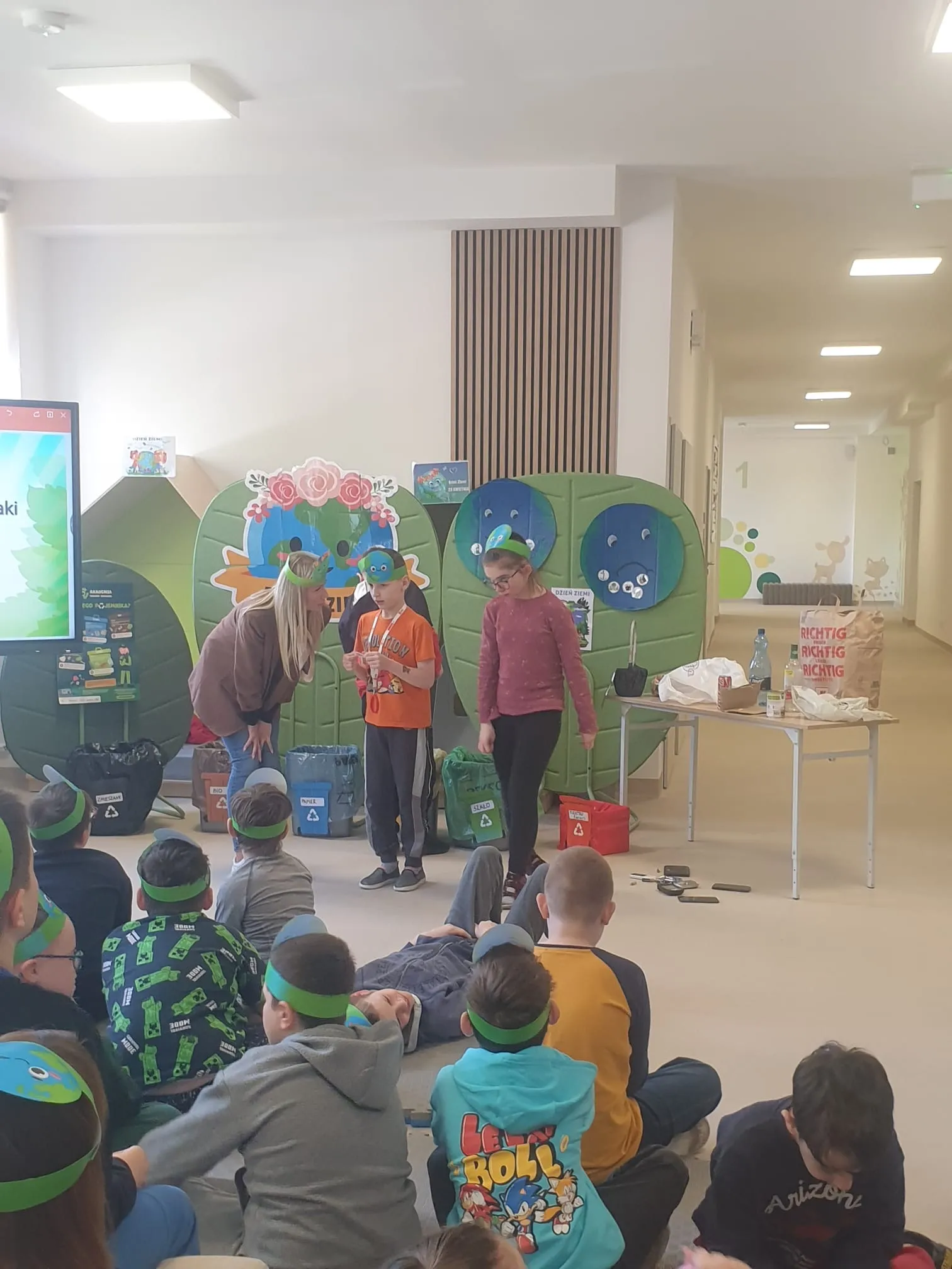 Na zdjęciu uczeń oraz uczennica, stojący przodem do siedzących na korytarzu  innych uczniów.  Do stojących uczniów pochyla się pani nauczycielka. W tle widoczne są koszę na odpady, dekoracje związane z Dniem Ziemi oraz tablica multimedialna.