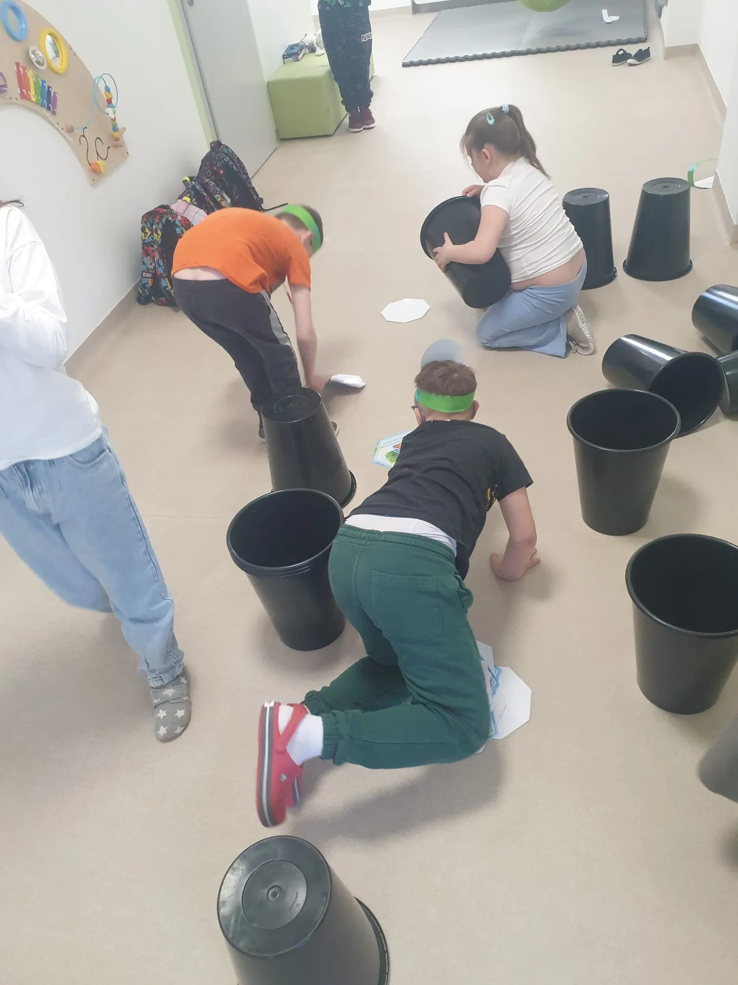 Na zdjęciu widoczni są uczniowie, którzy pochylają się przy czarnych wiaderkach, które w dużej ilości stoją lub lezą na korytarzu szkolnym. Na podłodze widoczne są też papierki