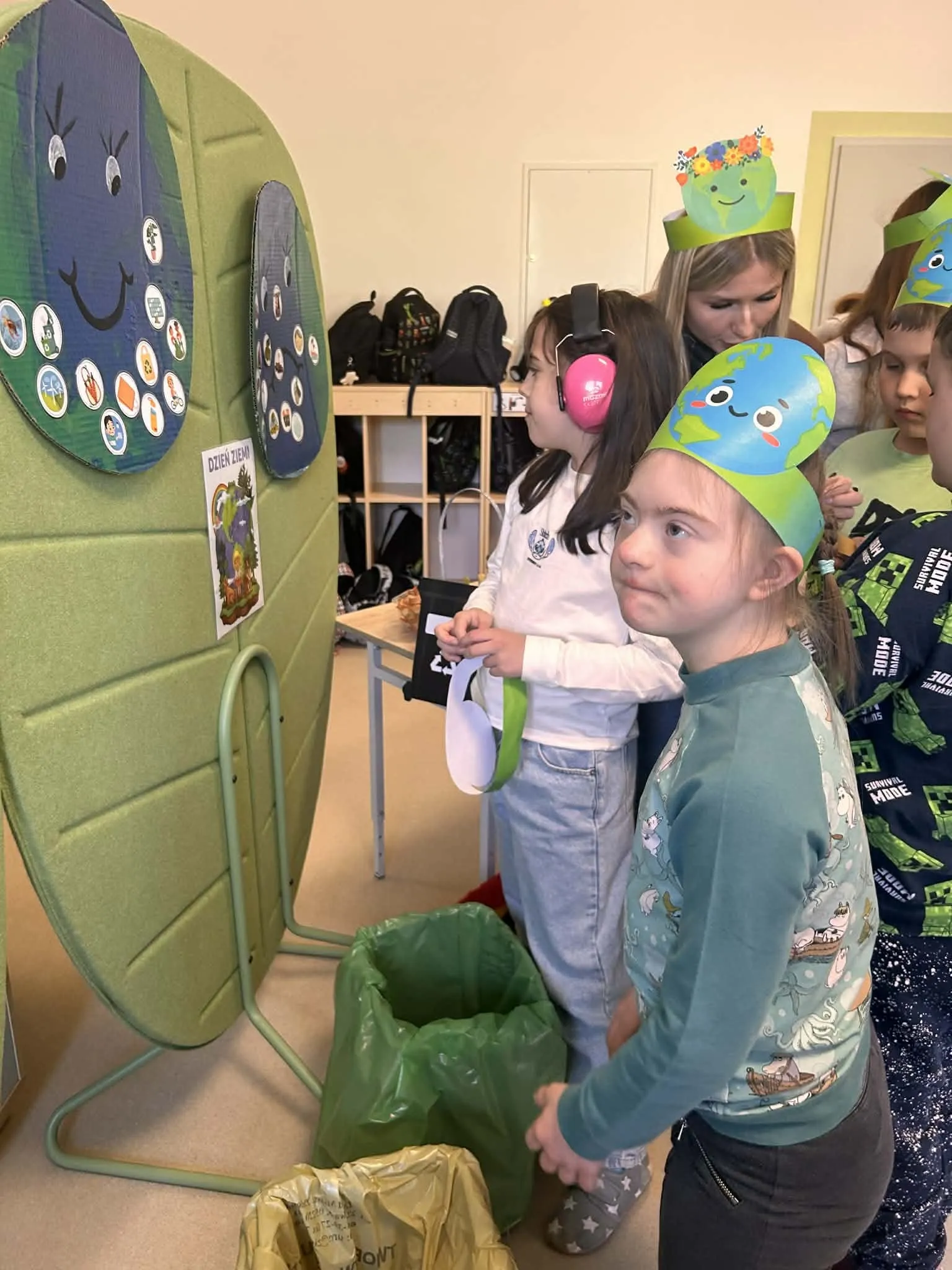 Na zdjęciu widoczna  uczennica , która stoi przed planszą  (umocowaną na zielonym tle) z naklejonymi na niej różnymi elementami.  Obok wisie podobna plansza. Przodem do niej stoją uczniowie oraz nauczycielka. /wszyscy oprócz jednej uczennicy mają czapeczki  z wizerunkiem kuli ziemskiej.  