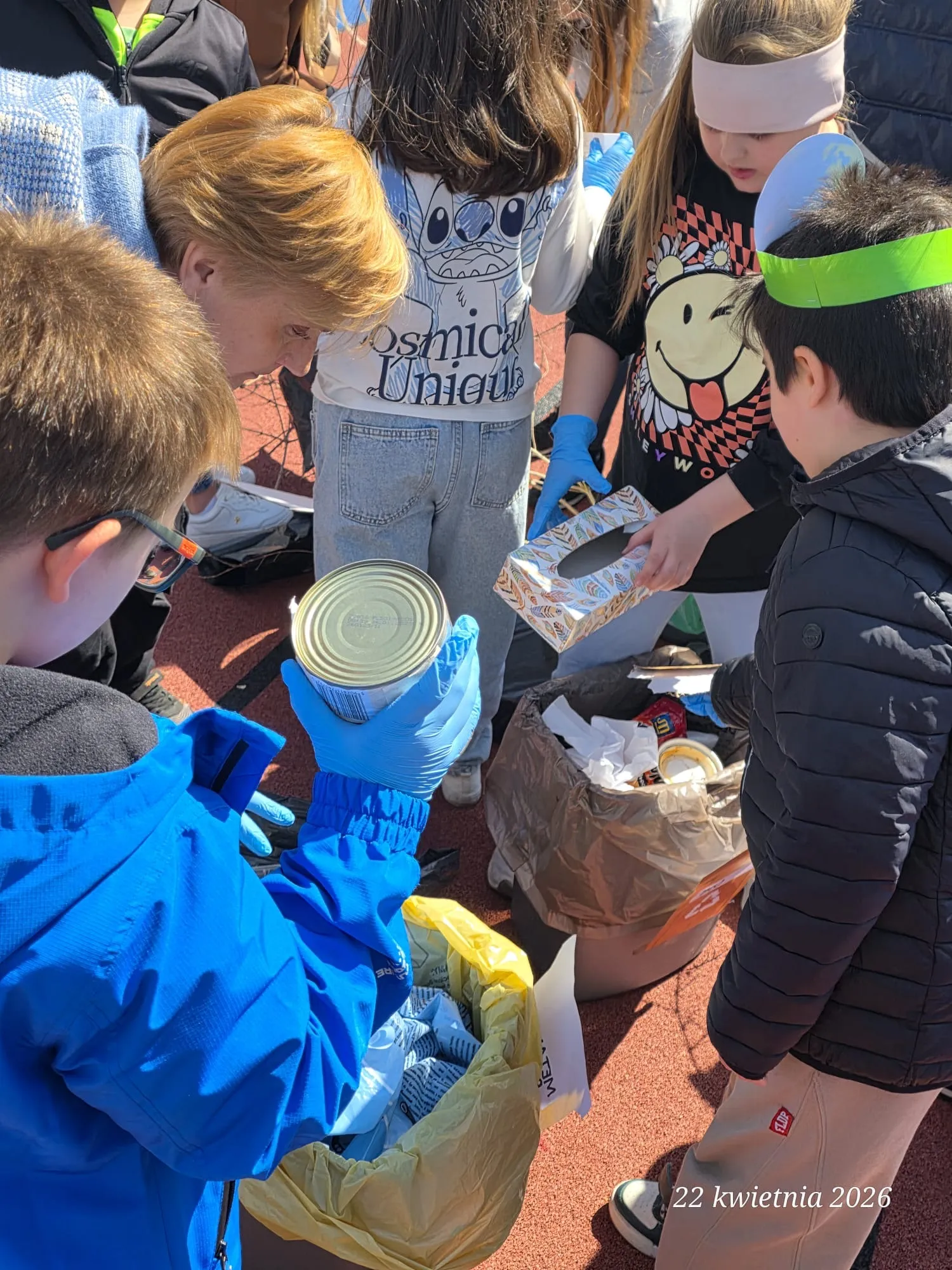 Na zdjęciu uczniowie oraz jedna nauczycielka. Znajdują się na boisku szkolnym  przy koszach na różne odpady. Dzieci trzymają w rękach różne odpady, które znajdą się w odpowiednich koszach.