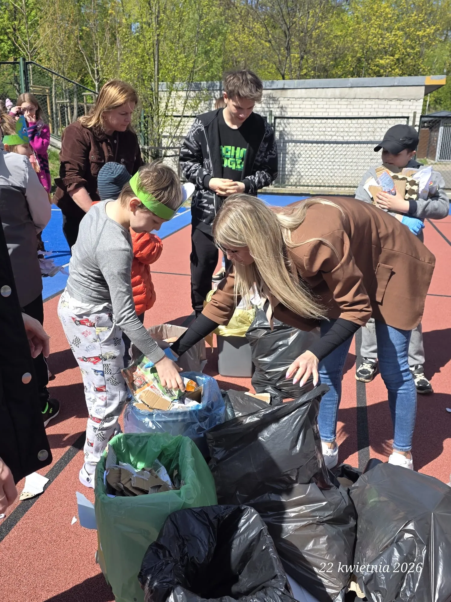 Na zdjęciu widoczni są uczniowie oraz nauczyciele, stojący w pobliżu  koszy na różne odpady.  Jeden z uczniów wkłada coś do kosza.