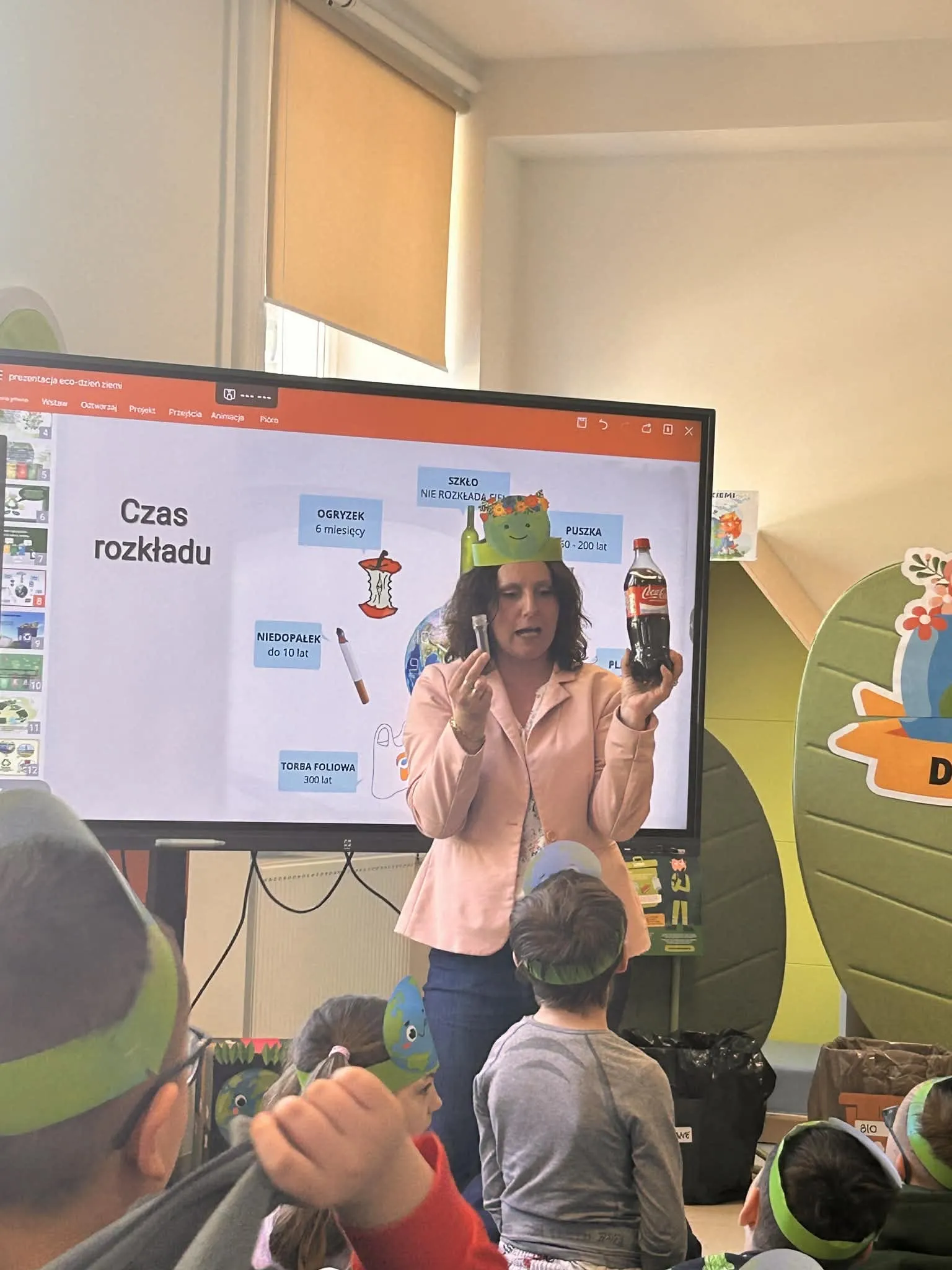 Na zdjęciu nauczycielka na tle tablicy interaktywnej ze slajdem o rozkładzie różnych materiałów. nauczycielka trzyma w ręku butelkę plastikową z colą i jakiś przedmiot.  Przed nauczycielką widoczni są siedzący na korytarzu uczniowie. 